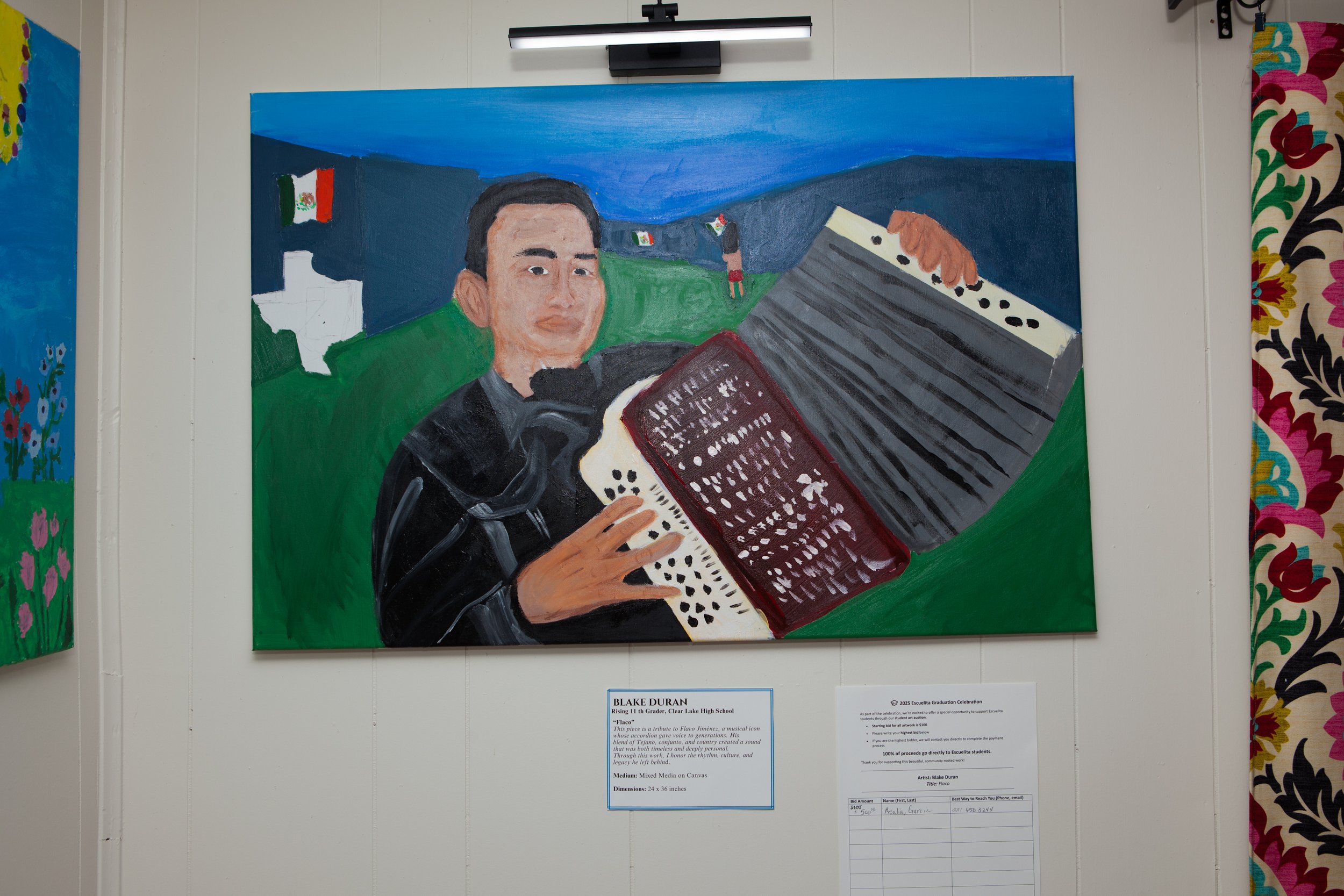 A painting of a young man playing an accordion, with a mountainous landscape in the background, flags, and small figures. The painting is displayed on a wall with a label below it.