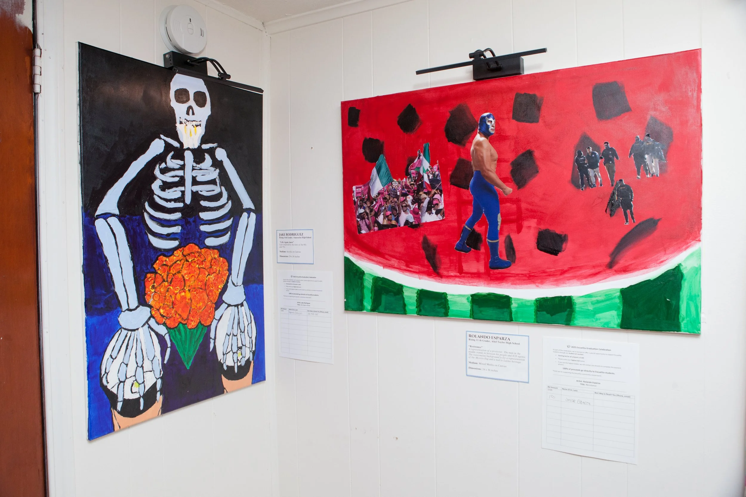 Art display in a gallery featuring a large painting of a skeleton holding an orange bouquet on the left, and a smaller collage of images and a figure wearing a lucha libre mask on the right.