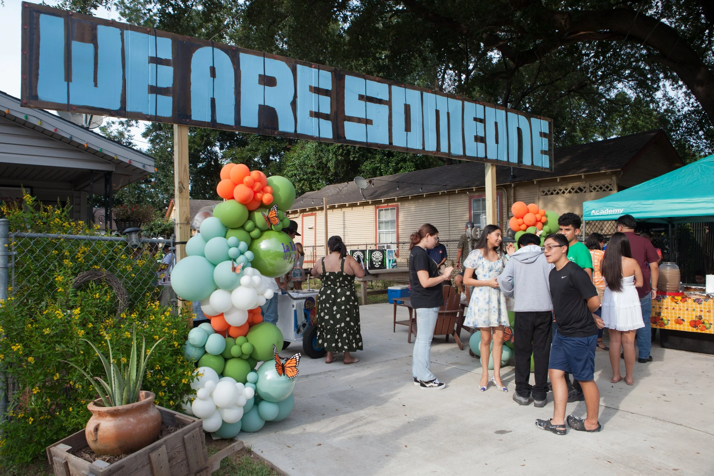 People gathered outdoors at a community event with a large sign that reads 'We are Someone.' There are balloons and a plant in a pot visible in the foreground, and several small groups of people are talking near tables and booths under a canopy.