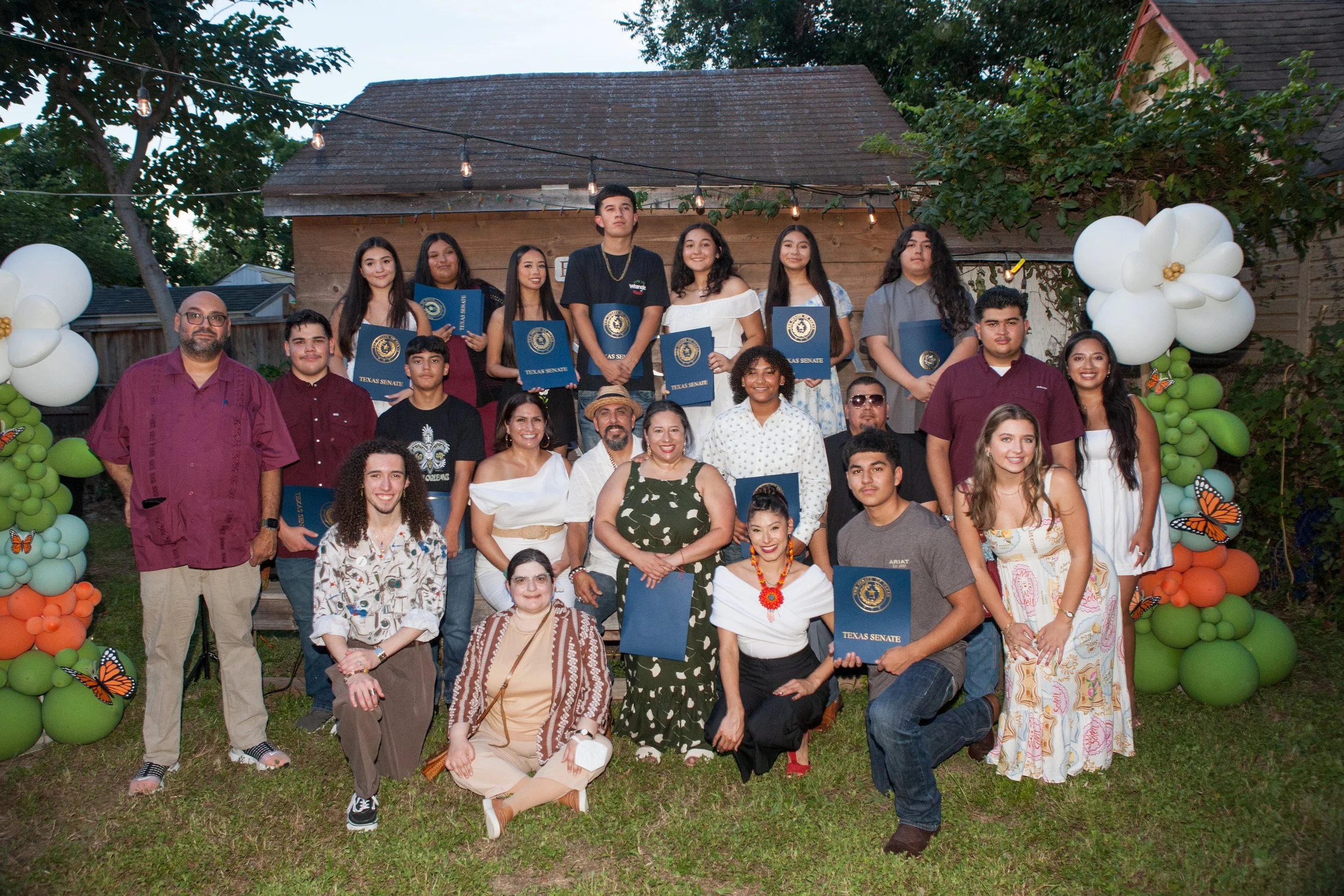 A group photo of young people and adults at an outdoor celebration, with some holding blue certificates labeled 'Texas Senate,' standing under string lights and surrounded by balloon decorations in orange, green, white, and black with butterfly desig
