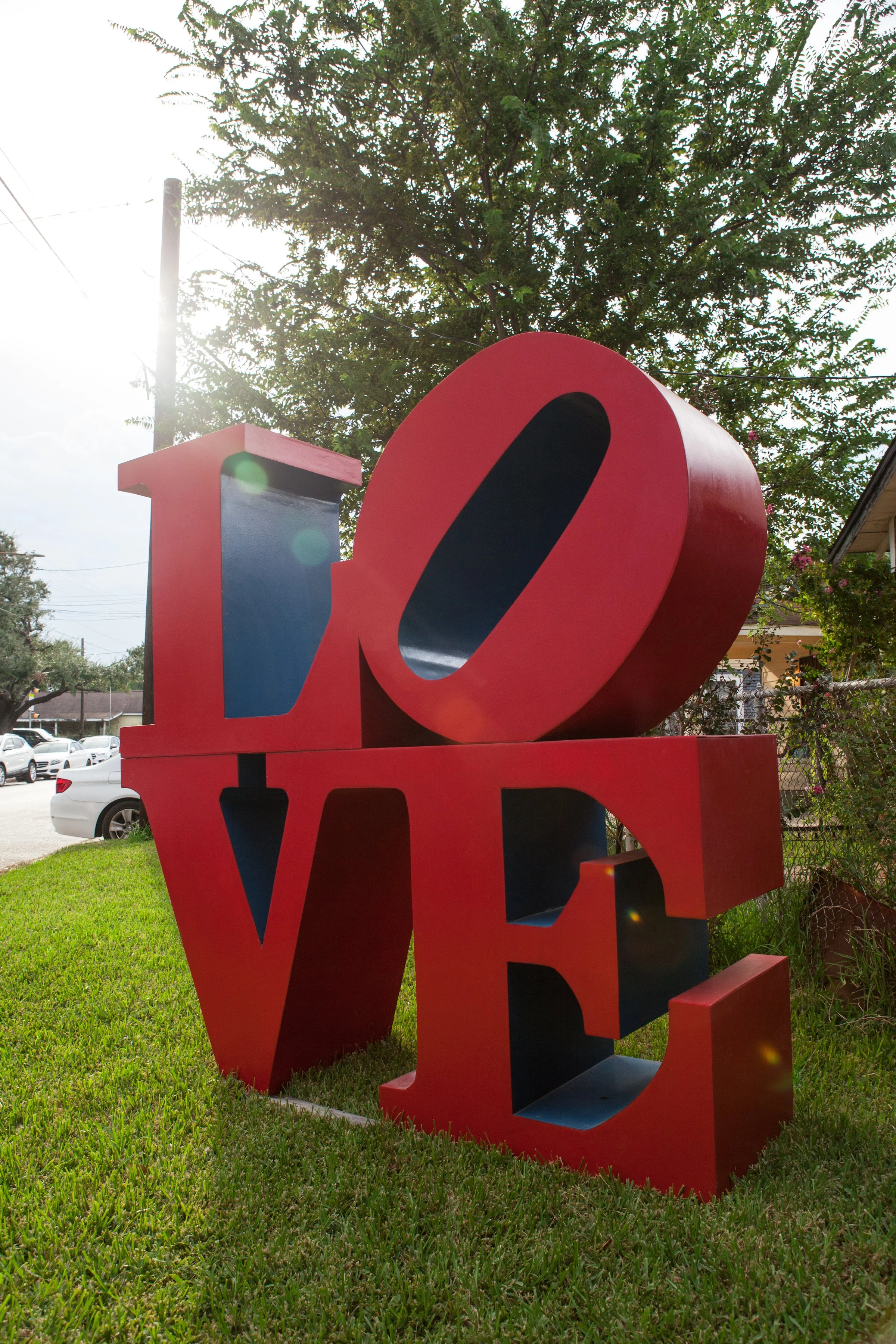 A large red and blue sculpture of the word "LOVE" outdoors on a grassy area with trees and a house in the background, sunlight creating lens flare.