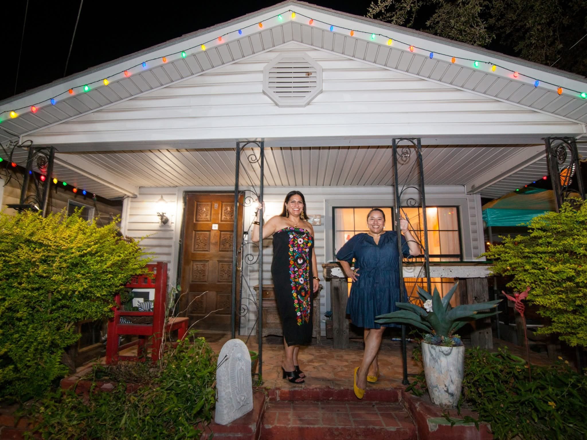 Two women standing on a porch decorated with colorful string lights at night, smiling and posing for the photo.