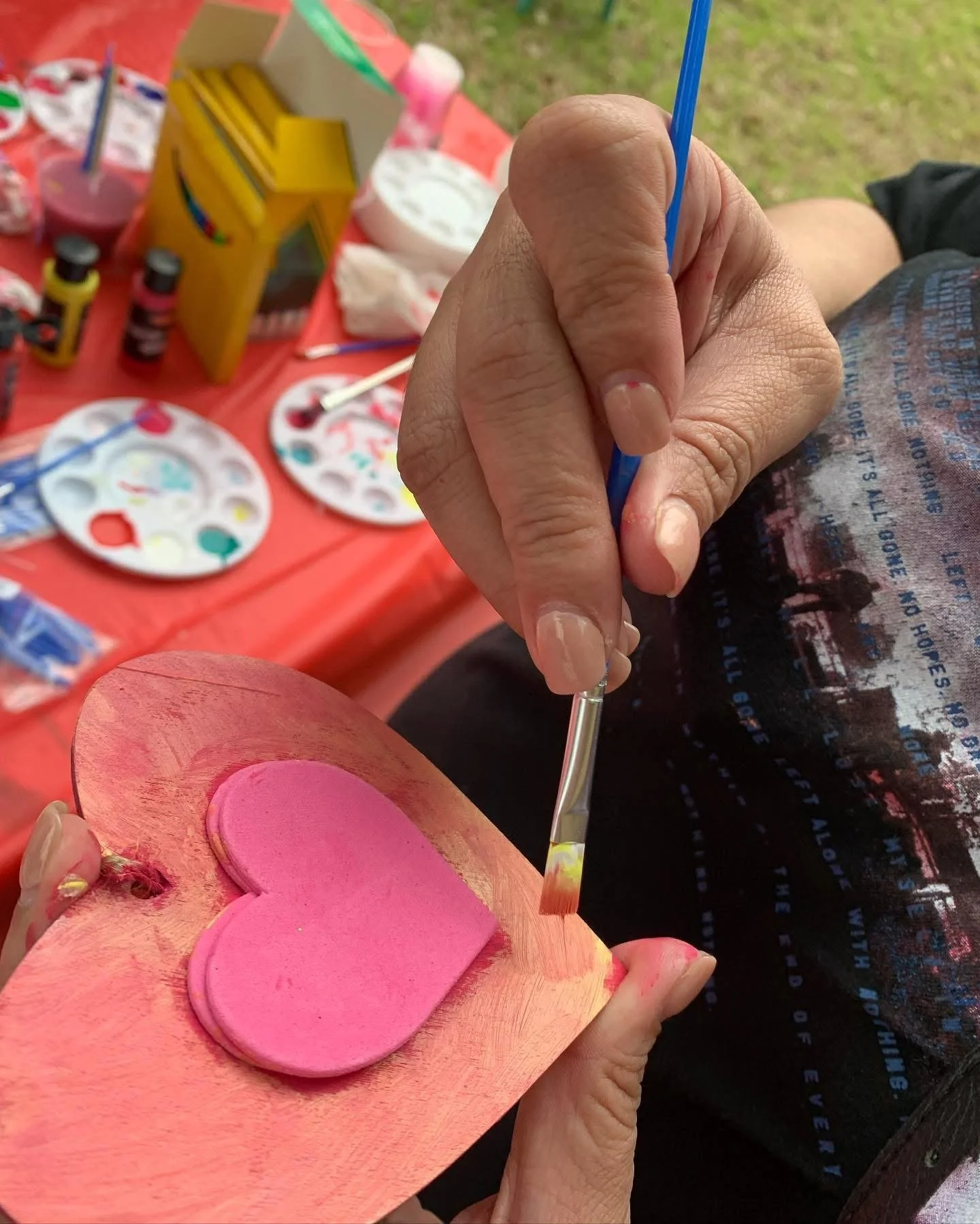 A person painting a pink heart on a wooden heart-shaped cutout using a small paintbrush. The background shows various paint supplies on a red tablecloth, including paint bottles, palettes, and a box of paintbrushes.