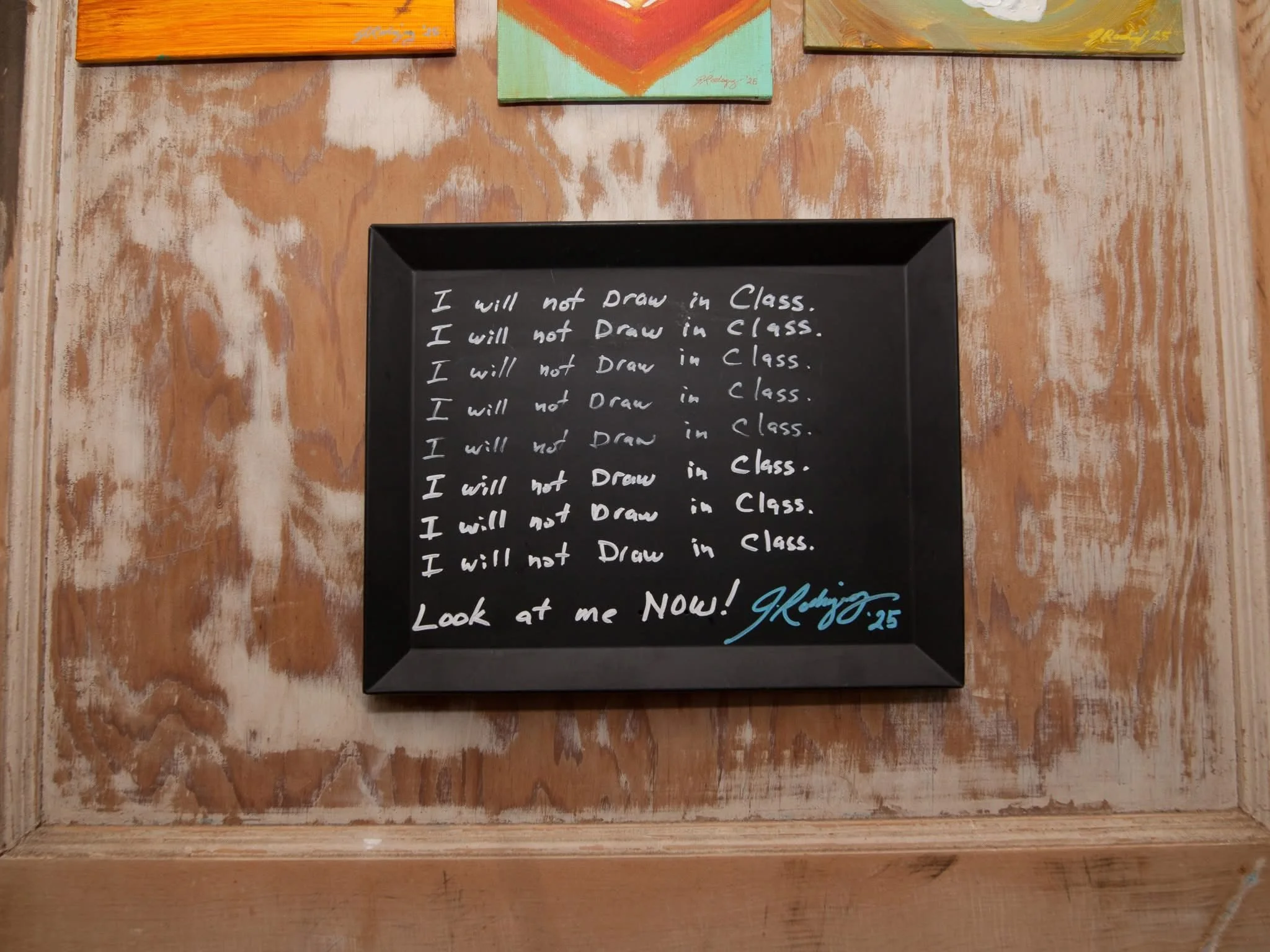 A blackboard with a message written in white chalk reading "I will not Draw in Class" repeated multiple times. The blackboard is placed on a wooden surface with a watercolor background. The earliest message ends with "Look at me NOW!" and is signed w