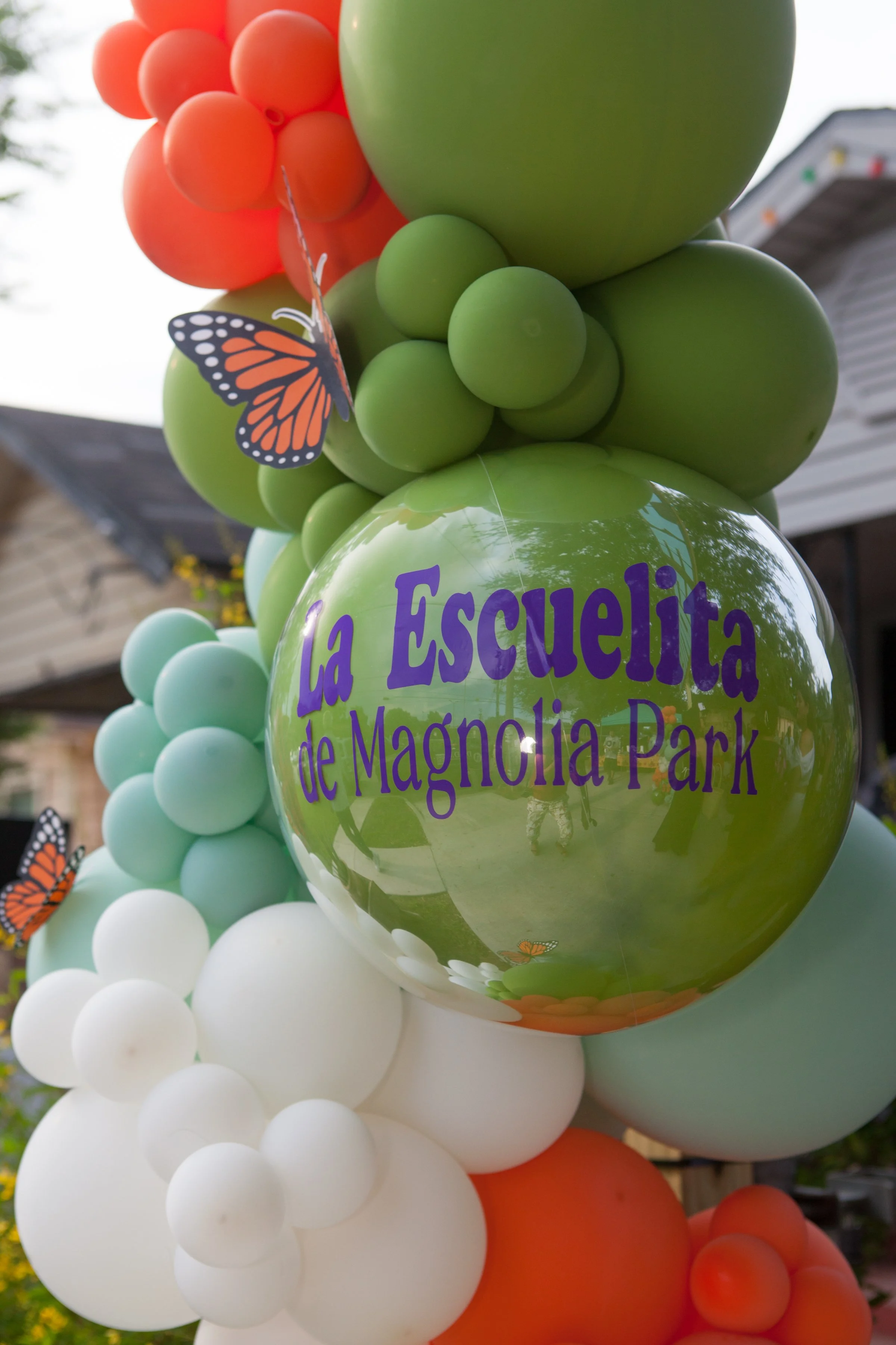 Colorful balloons in orange, green, white, and light blue, with a butterfly decoration, celebrating La Escuelita de Magnolia Park.
