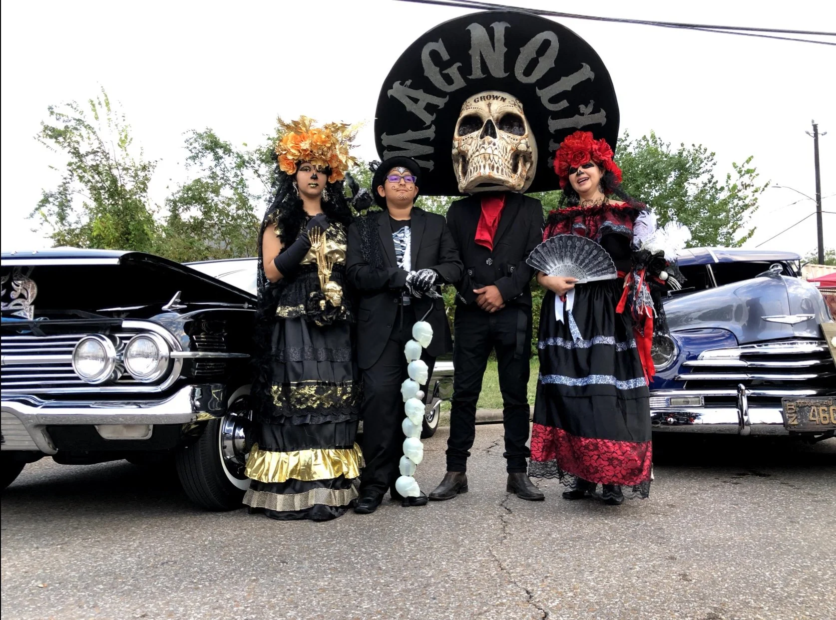 Four people dressed in traditional Day of the Dead costumes standing in front of vintage cars and a large black sign with a skull and the word "MEXICO" on it.