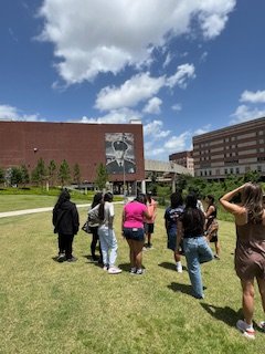 A group of people standing on a grassy area in front of a brick building with a large mural of a person's face on it, under a partly cloudy sky.