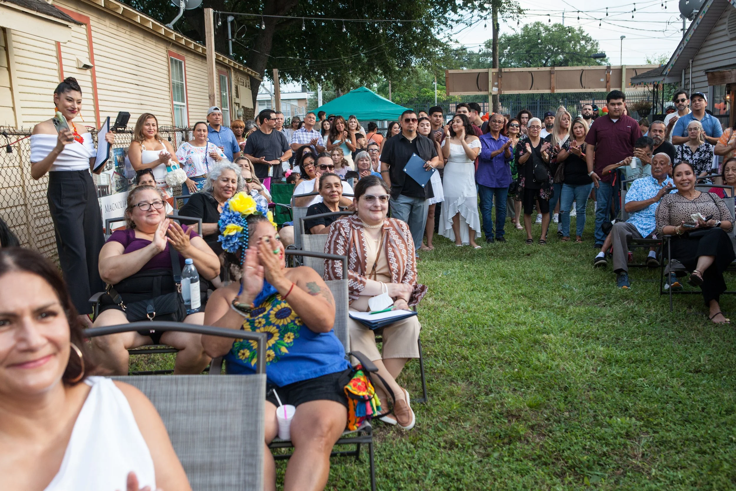 A diverse group of people gathered outdoors in a backyard, attending a social event, with some seated and others standing, and a woman in the foreground smiling and giving a thumbs up.