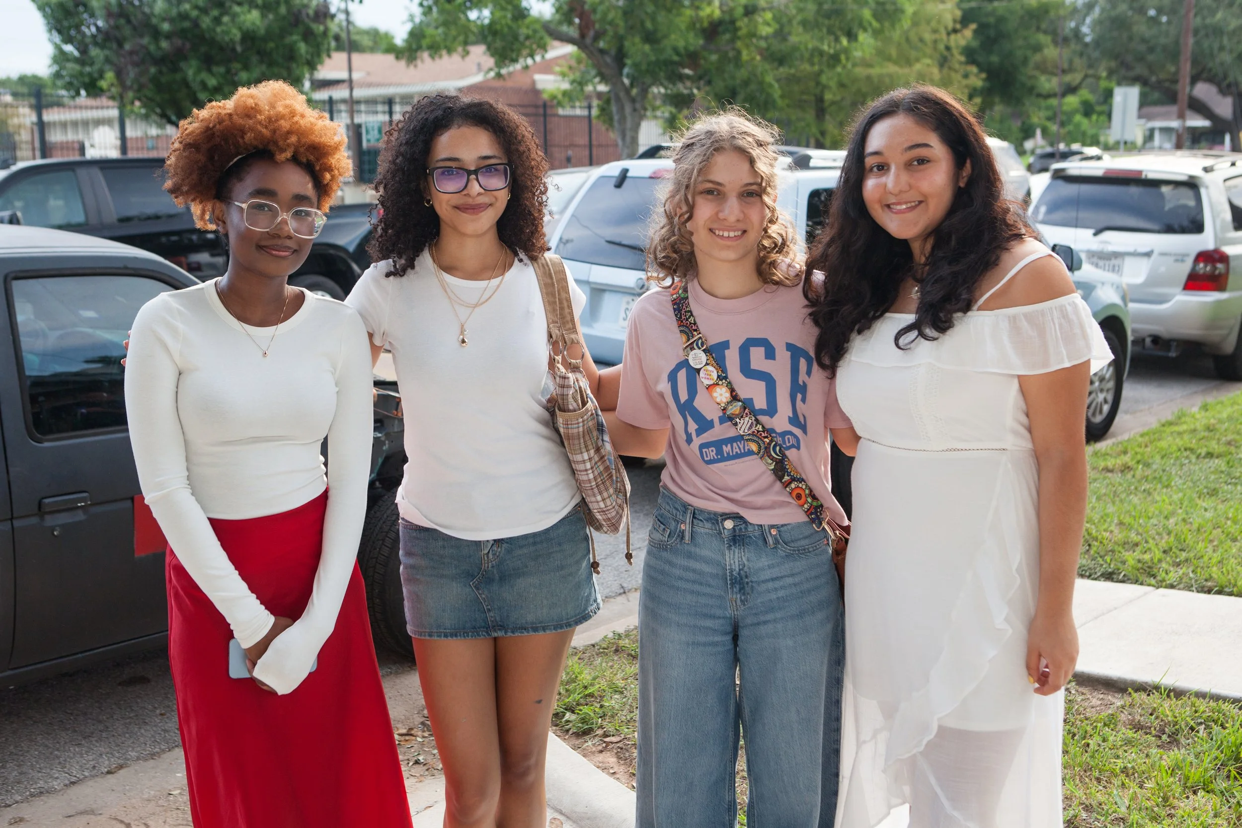 Four young women standing together outdoors, smiling, with cars and trees in the background.