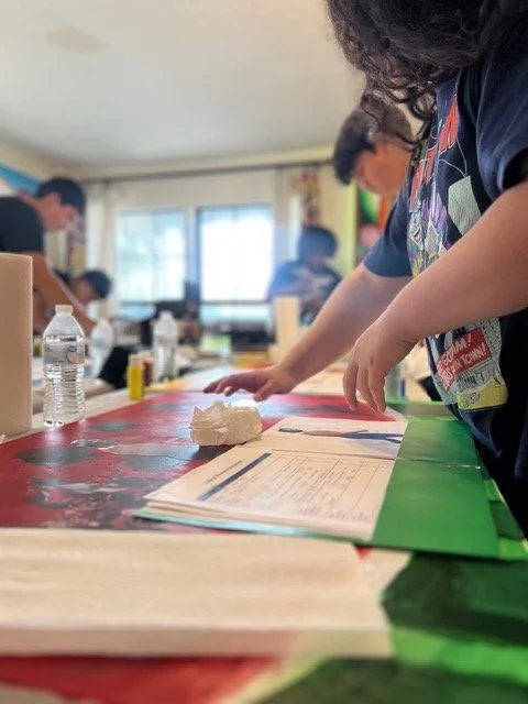 Students working at a table in a classroom, with papers, a water bottle, and a crumpled tissue visible.