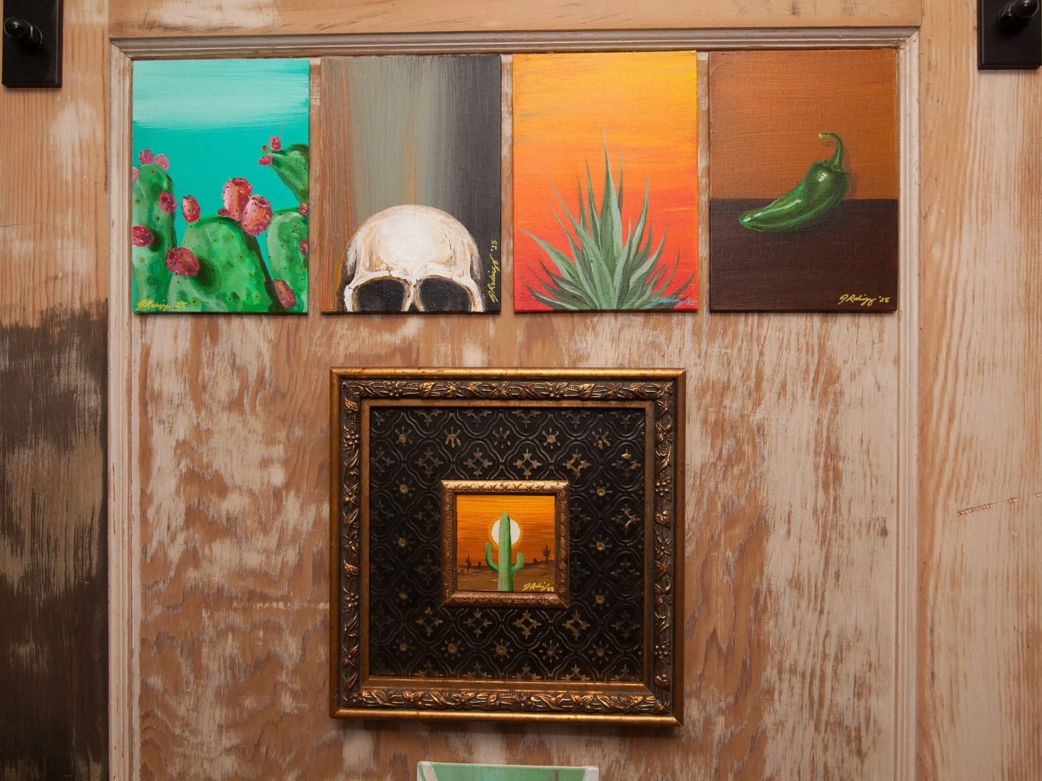Five paintings hang on a wooden wall. The first depicts prickly pear cacti with pink fruit against a blue background. The second shows a human skull on a neutral background. The third features an orange background with a green plant. The fourth illus