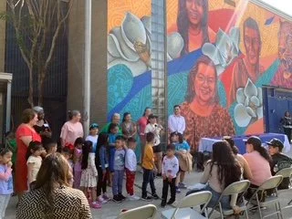 Group of children and adults gathered outdoors in front of a colorful mural with portraits and cartoon-like animals, some seated and others standing