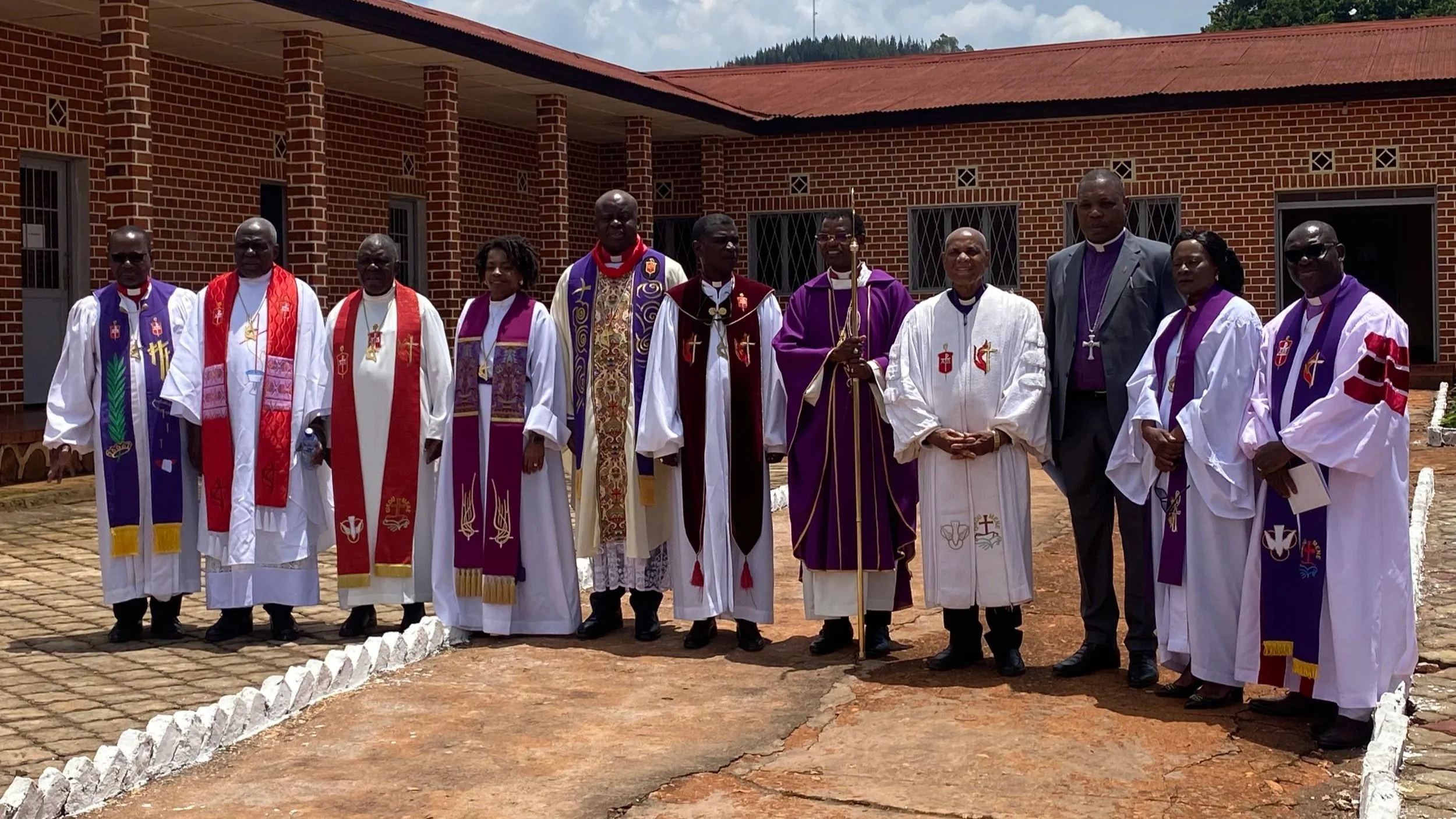 A group of United Methodist bishops and clergy stand outdoors in front of a brick building in Burundi, wearing liturgical robes in white, red, and purple.