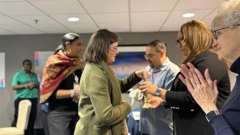 DEI roundtable participants sharing in communion, with one person offering bread while others receive, as participants nearby watch