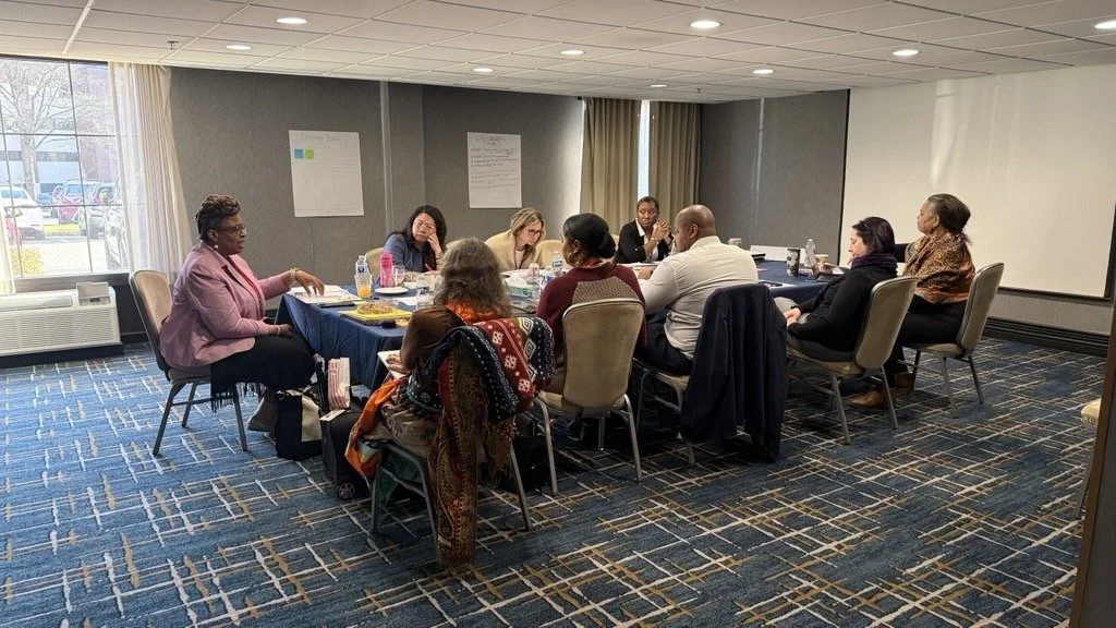 a small group of roundtable participants sit around a table in a meeting room engaged in discussion, with notes and papers visible as one person speaks and others listen.