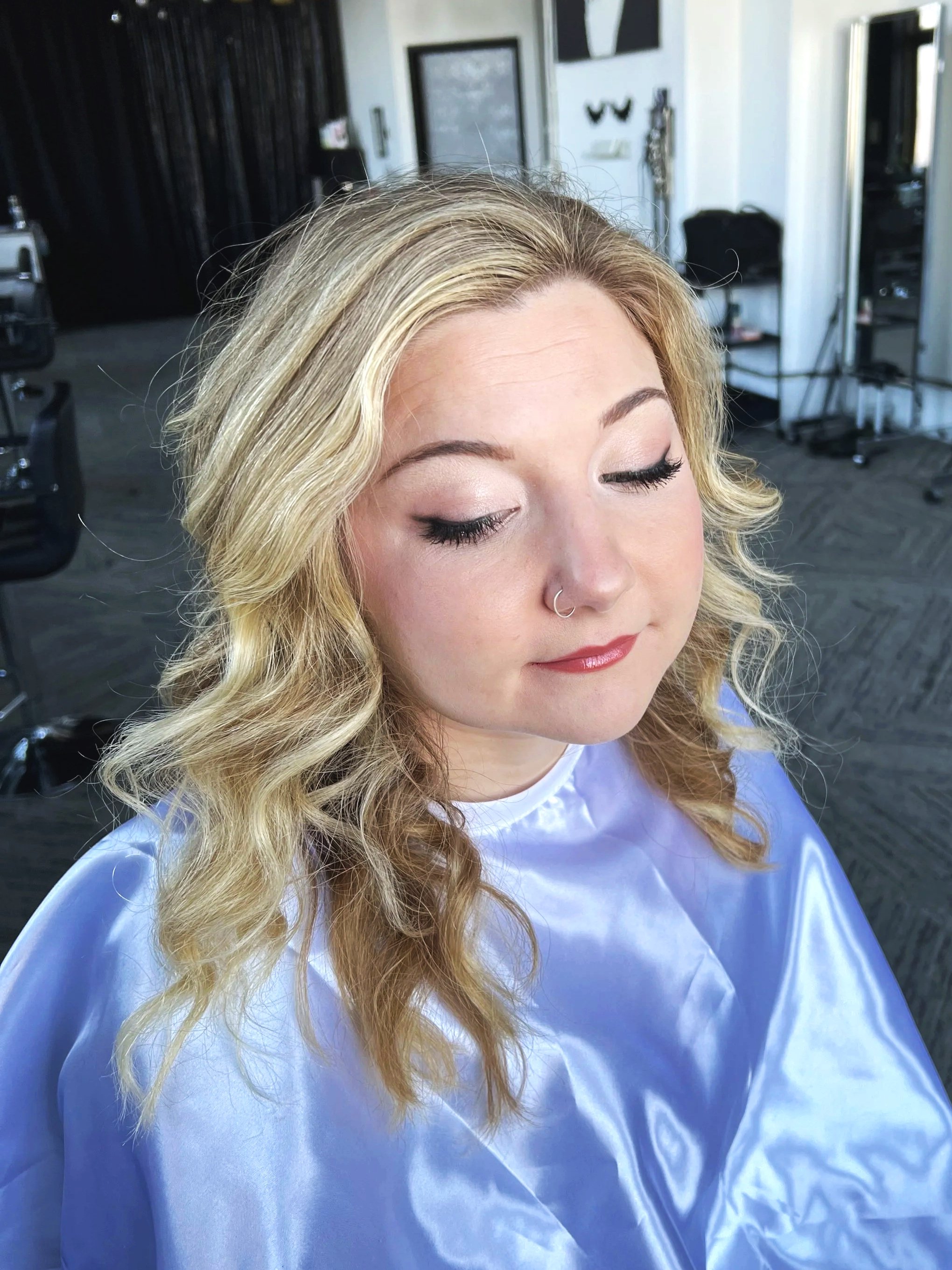 A woman with blonde, wavy hair and closed eyes, wearing a blue salon cape in a hair salon with black and white decor and styling tools in the background.