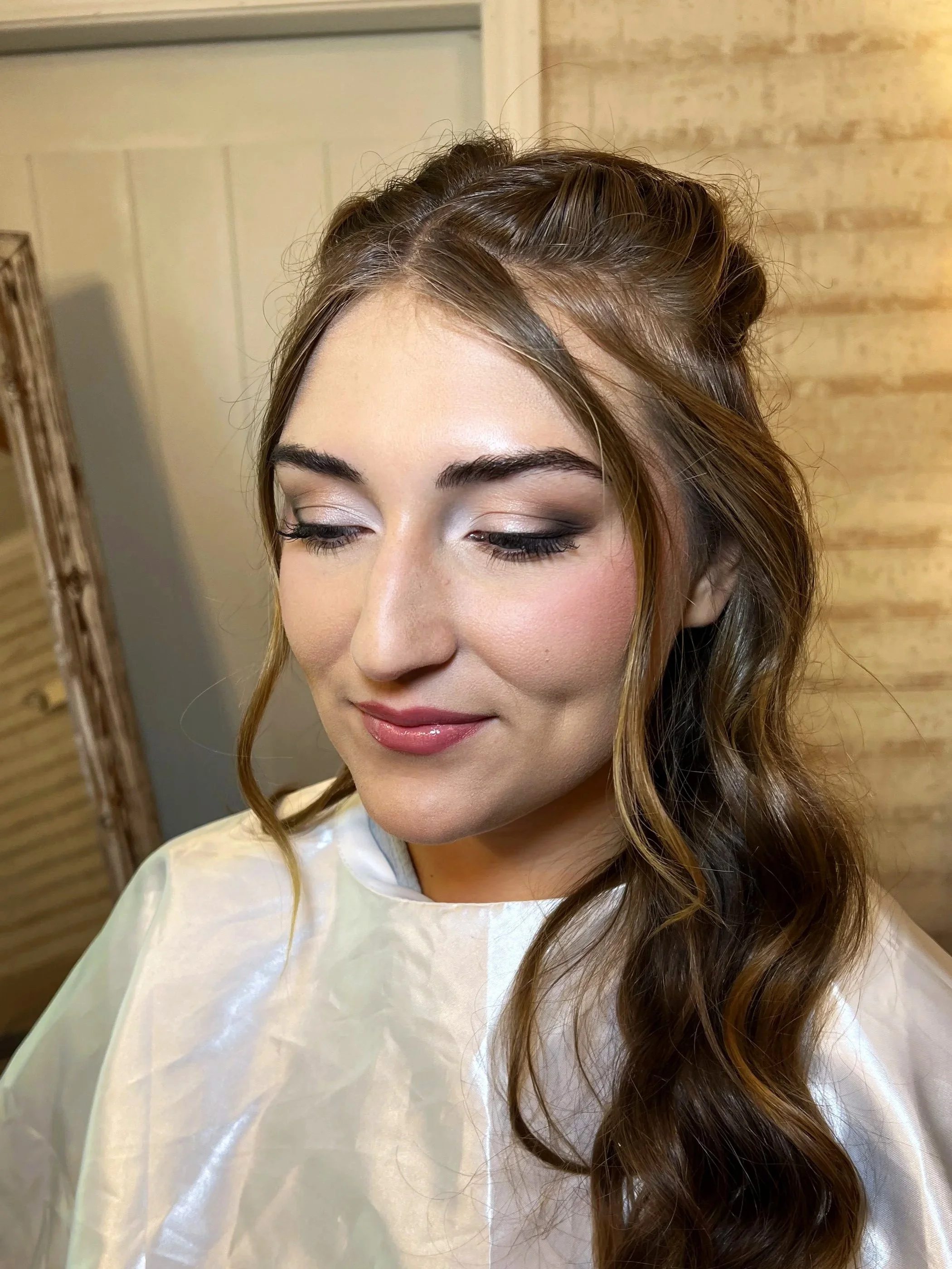 A woman with long brown hair styled in loose waves, wearing makeup with light eyeshadow, winged eyeliner, mascara, blush, and glossy lipstick, is smiling with her eyes closed, sitting in a salon or studio with a wooden wall and mirror in the background.