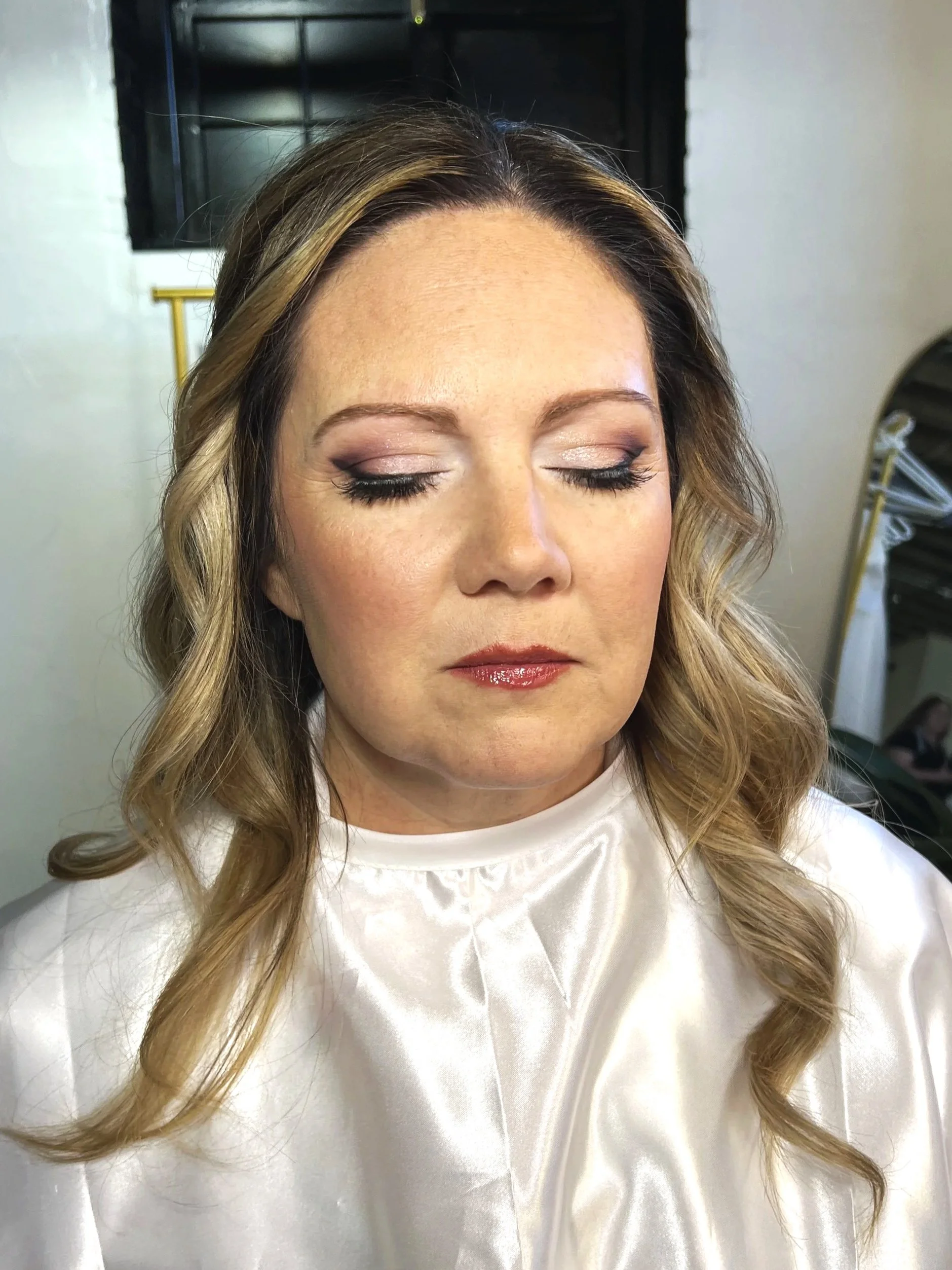 A woman with styled blonde hair, wearing a white satin-like top, with her eyes closed and makeup including eyeshadow, eyeliner, mascara, and lipstick.
