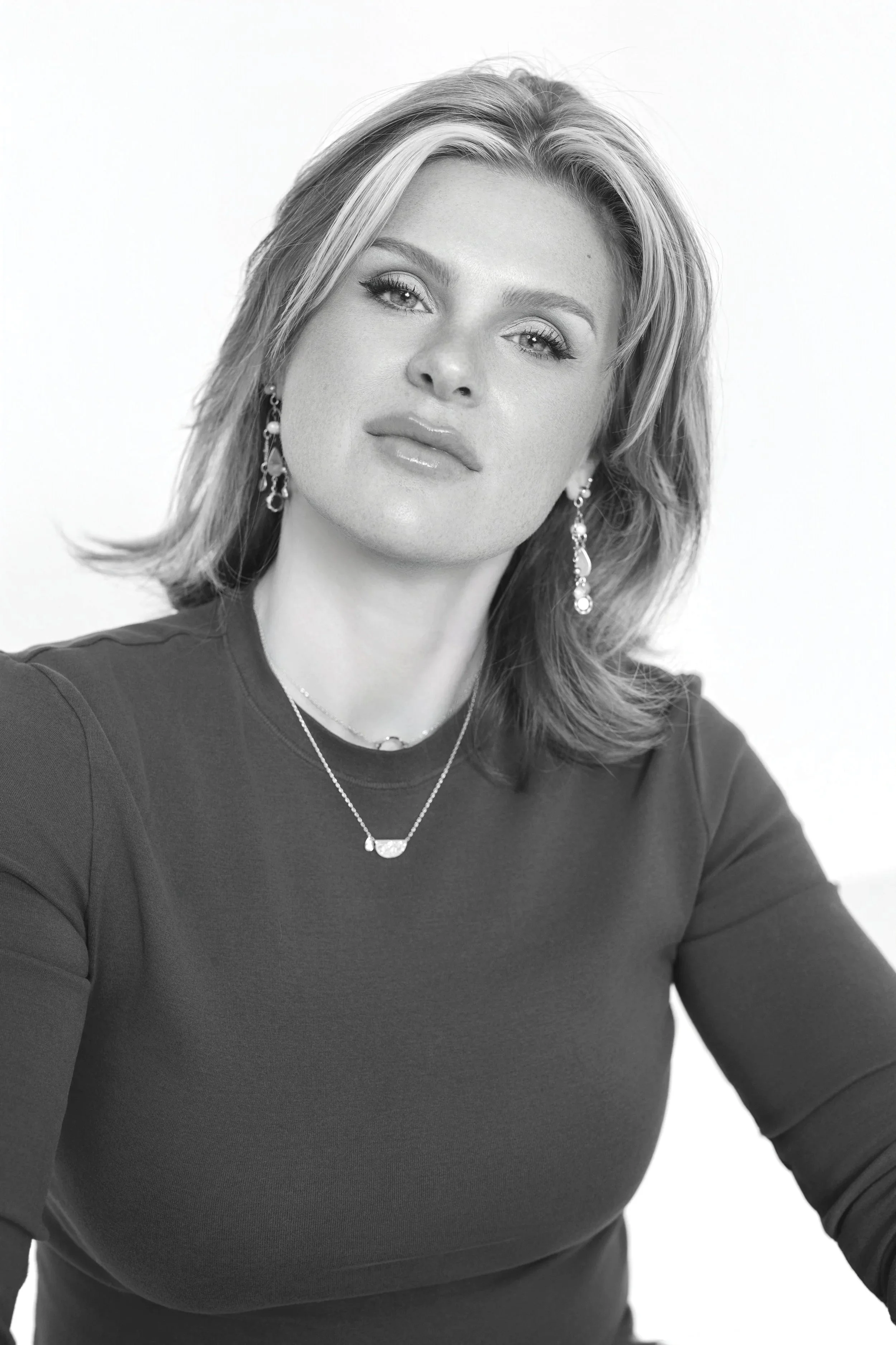 Black and white portrait of a woman with shoulder-length hair and light eyes, wearing earrings and layered necklaces, in a relaxed pose against a plain background.