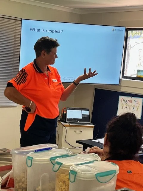 A woman in an orange shirt giving a presentation on respect in a classroom or training room, with a large screen behind her displaying the question 'What is respect?' and a laptop on a desk nearby.