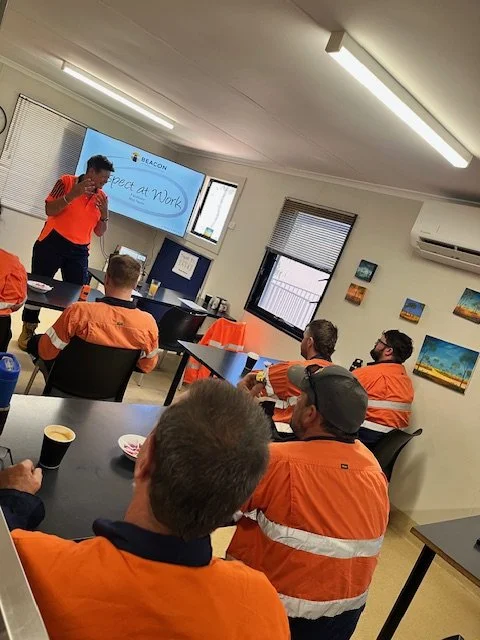 People wearing orange safety vests attending a presentation in a conference room. The presenter is standing near a screen that displays the words 'Respect at Work.'