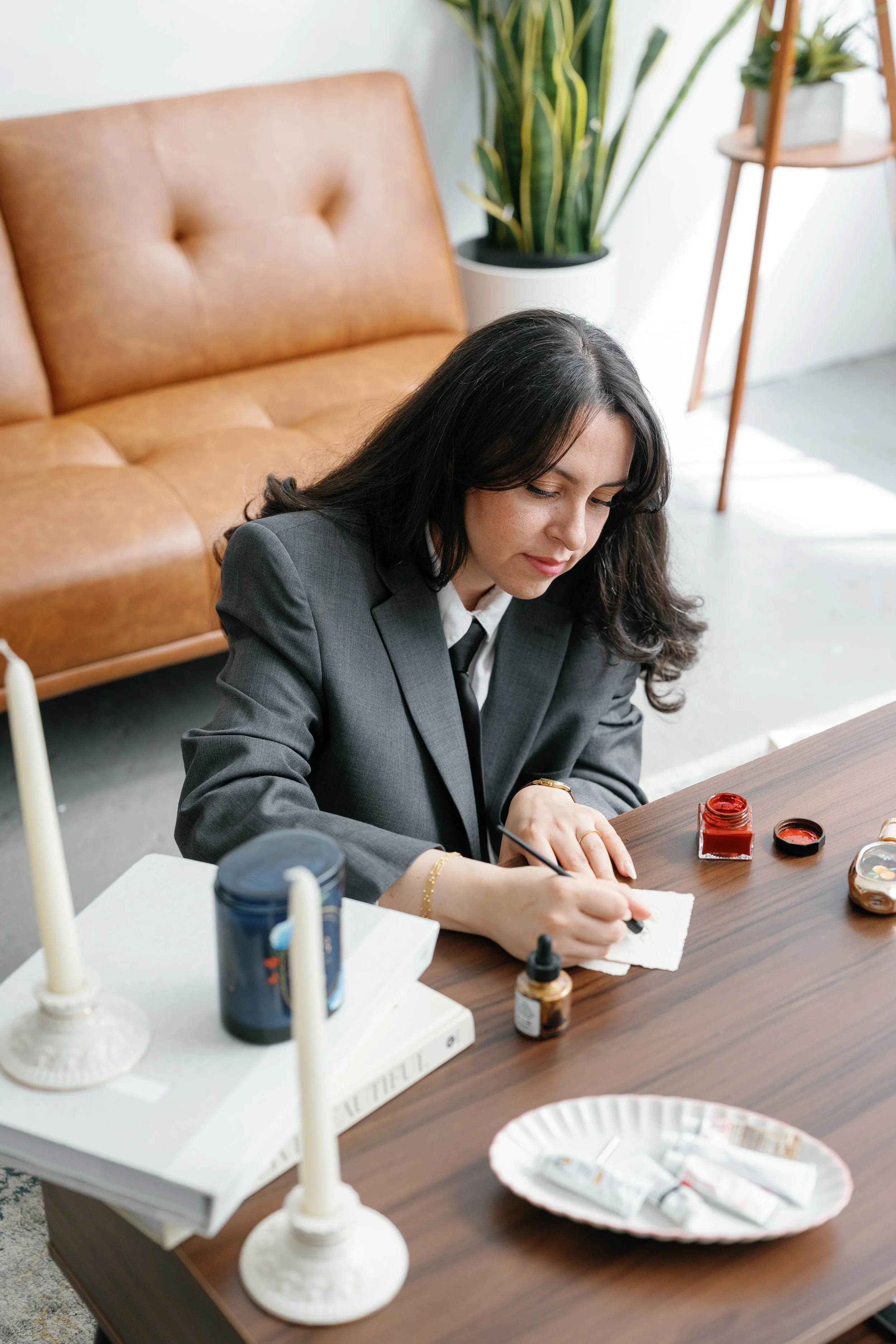 A woman in a gray blazer is sitting at a table, painting or decorating a small object with red paint. The table has various art supplies, including small bottles of paint, a paper towel, and a tray with white packages. In the background, there is a b