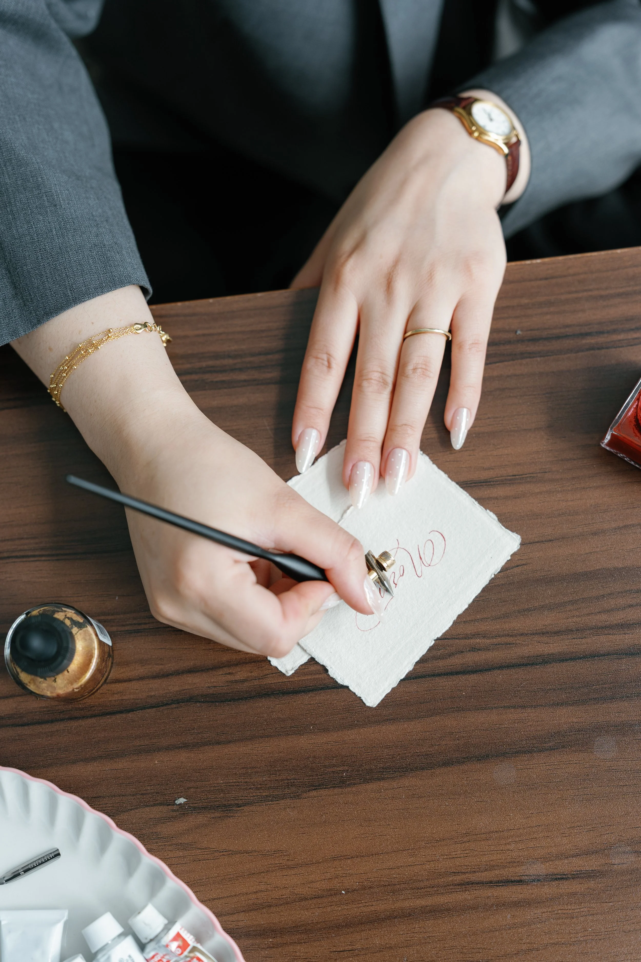 Close-up of a person's hand writing calligraphy with a paintbrush on a piece of paper, with various jewelry including a gold bracelet, a watch, and a ring visible on their wrist and finger. The person is sitting at a wooden table with art supplies in