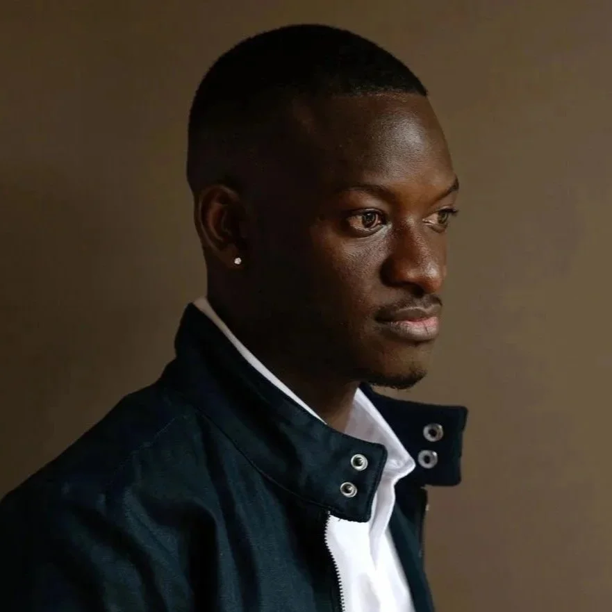 A young black man with a short haircut and stud earrings, wearing a dark jacket over a white shirt, looking to the right, against a plain background.