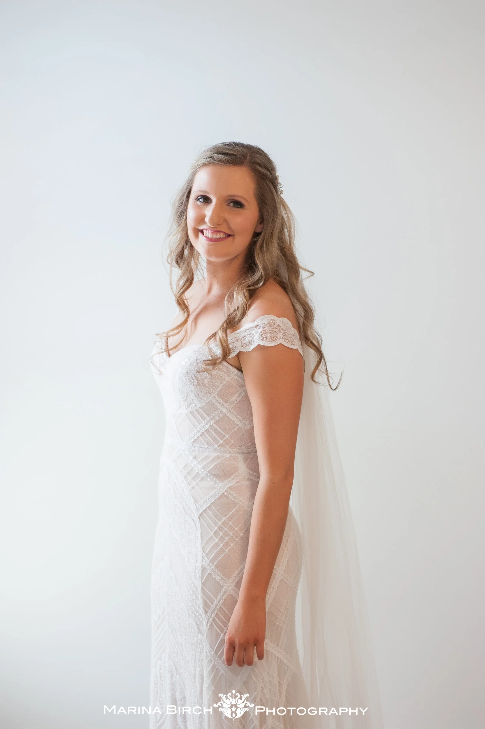 A woman with long wavy blonde hair in a white lace off-shoulder dress, smiling at the camera against a plain white background.