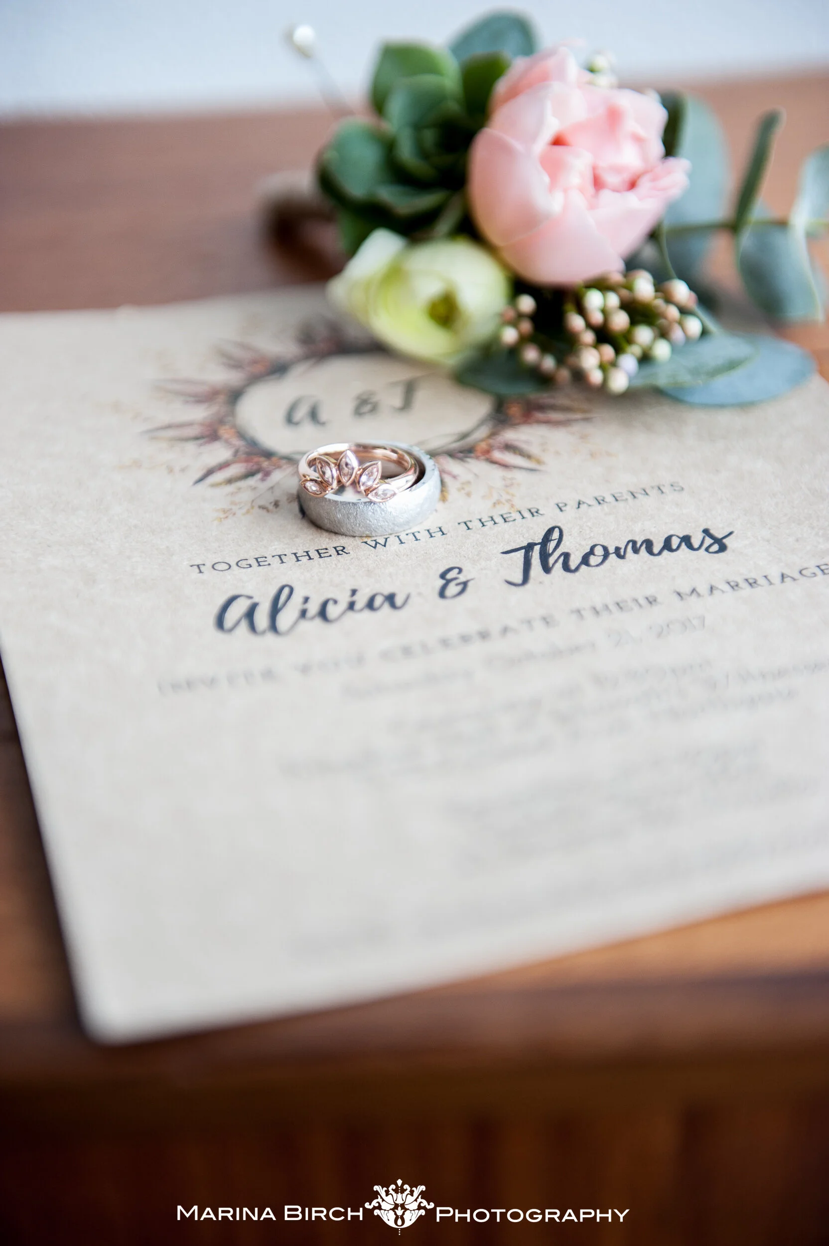 A wedding invitation with rings placed on top, accompanied by a bouquet of pink and cream flowers and green foliage. The invitation mentions Alicia and Thomas and features decorative text and design.
