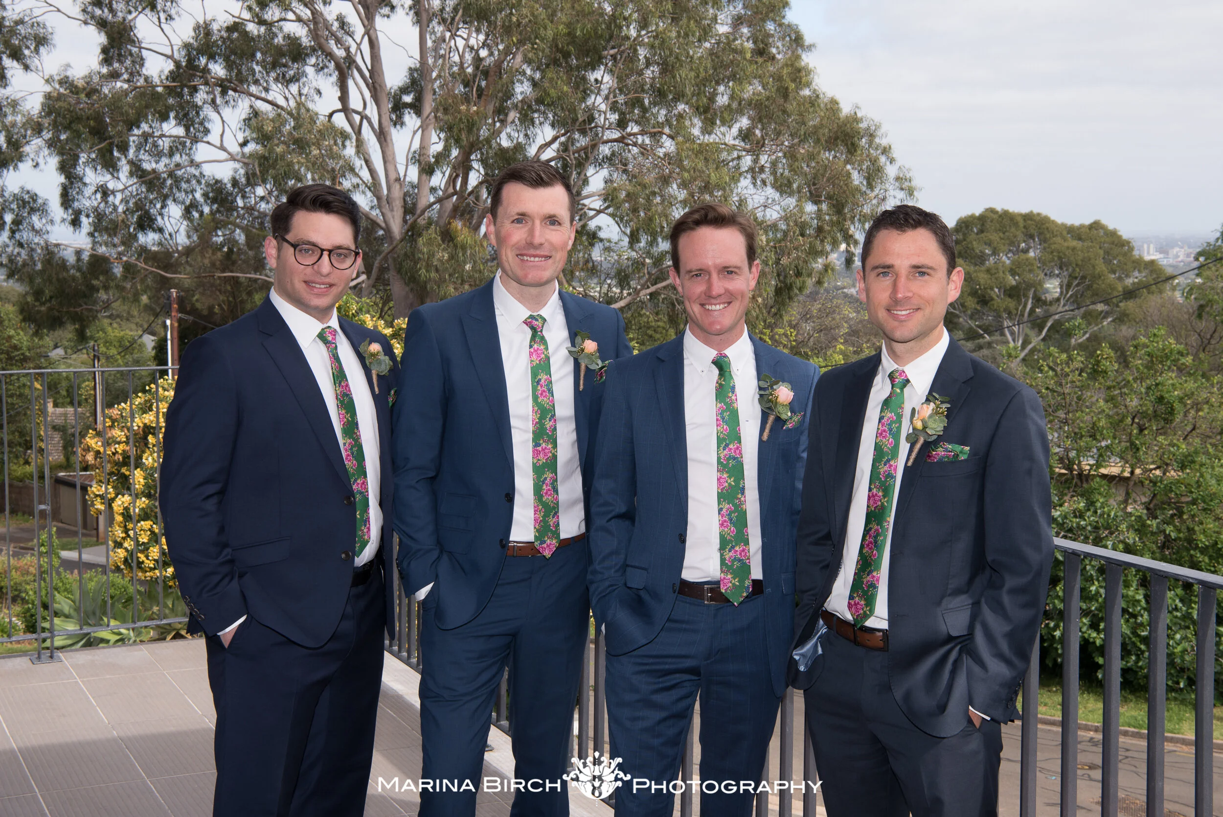 Four men in suits with floral ties and boutonnieres standing outdoors on a balcony, with trees and a cloudy sky in the background.
