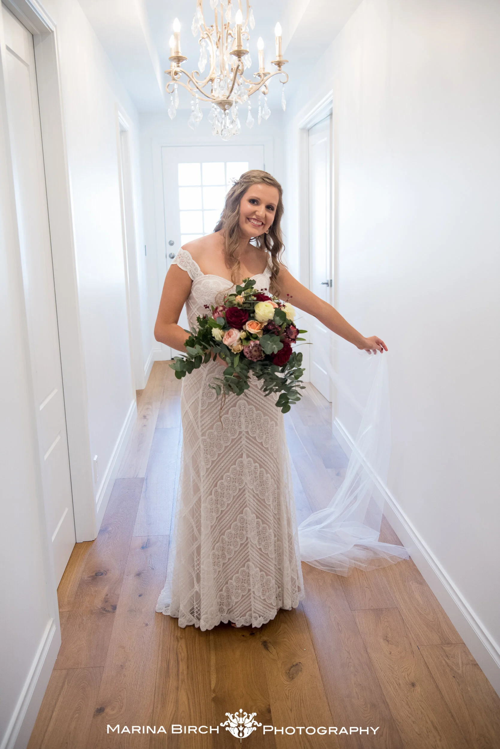 A smiling bride in a white lace wedding dress holding a bouquet of mixed flowers, standing in a bright hallway with a chandelier overhead and wooden flooring.