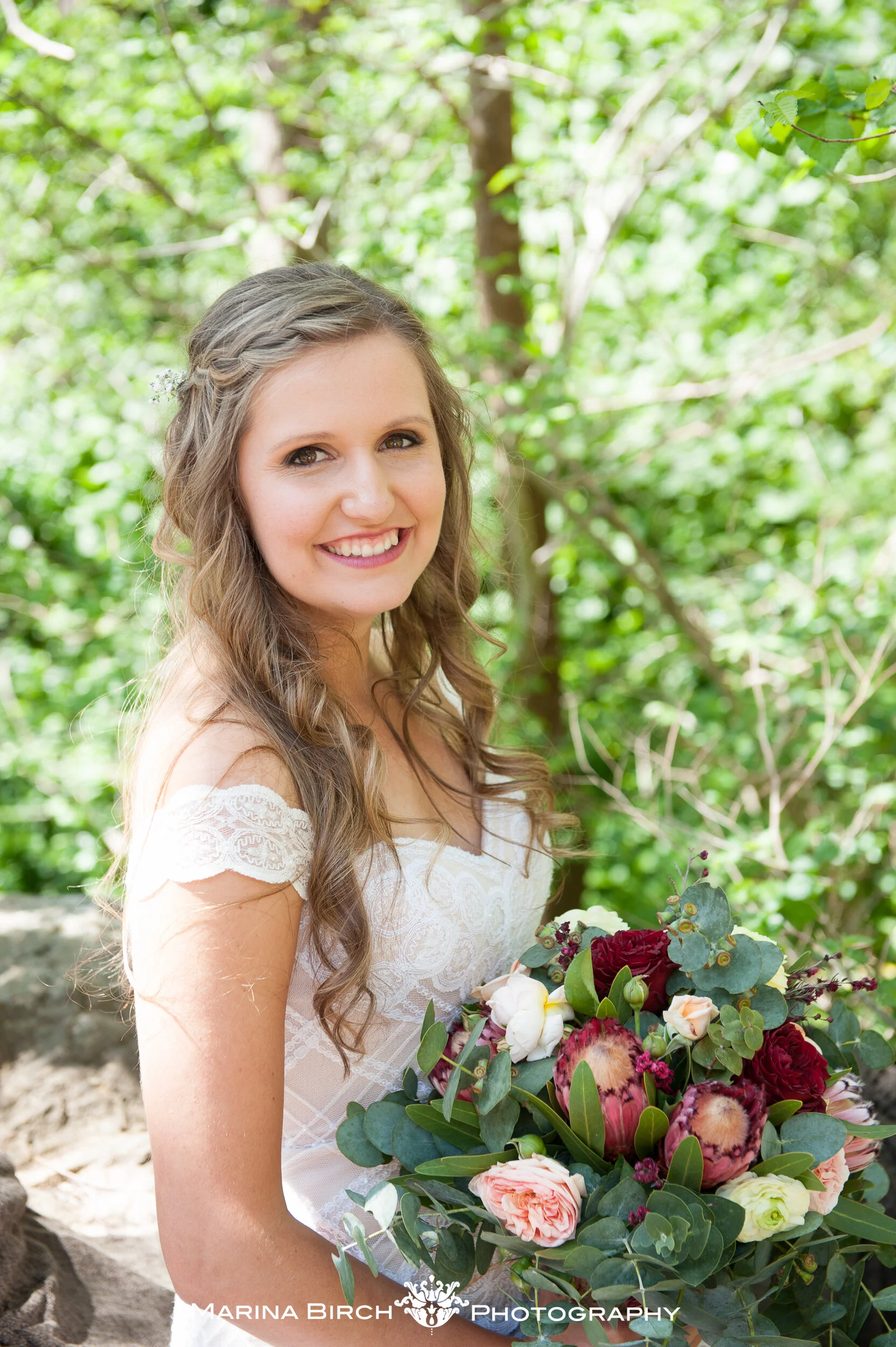 A smiling woman in a white lace dress holding a bouquet of red, pink, and white flowers and greenery, standing outdoors with lush green trees in the background.