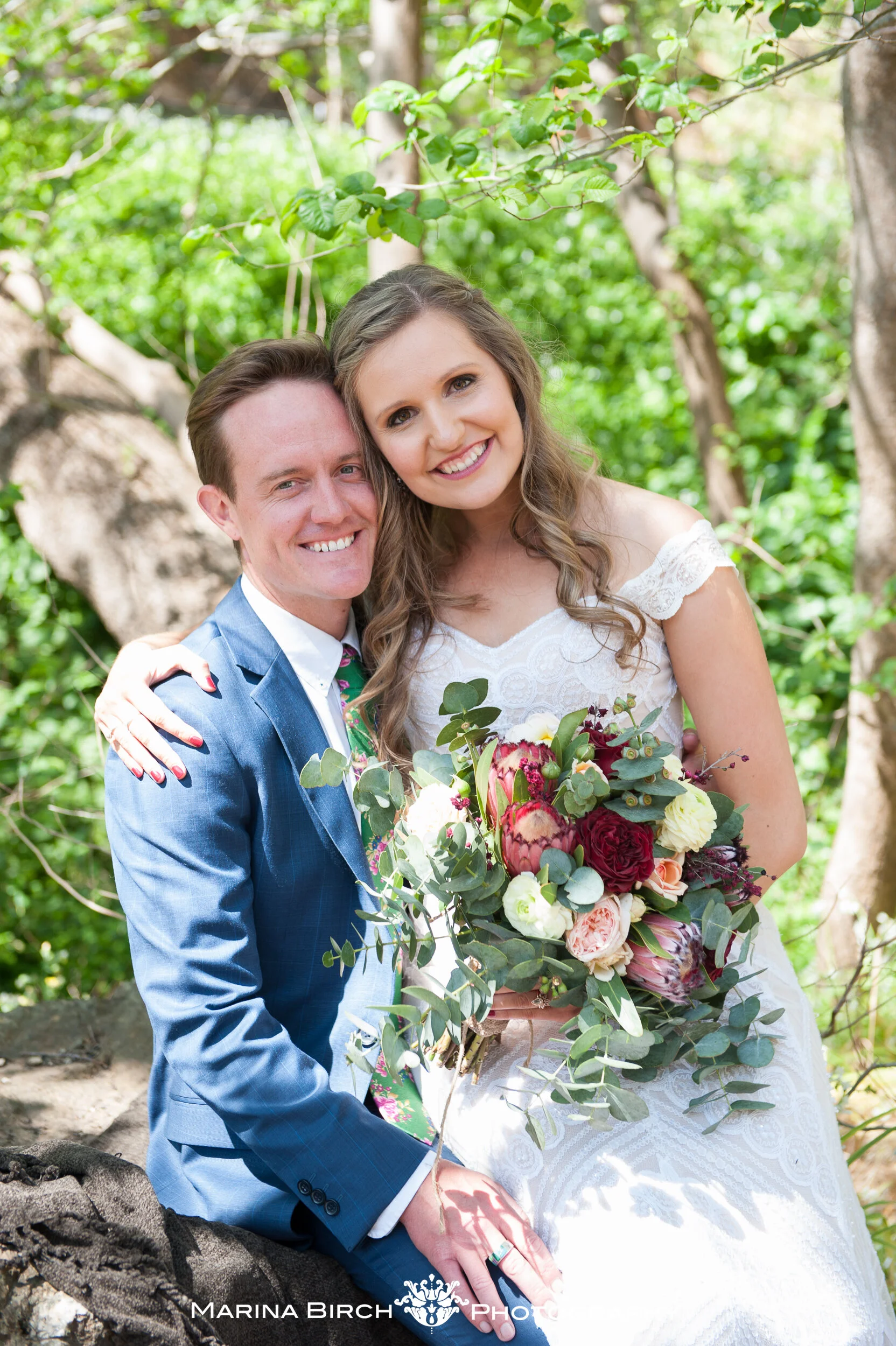 A couple in wedding attire smiling outdoors, surrounded by green trees. The woman holds a bouquet of flowers with roses, proteas, and greenery, and the man wears a blue suit with a colorful tie.