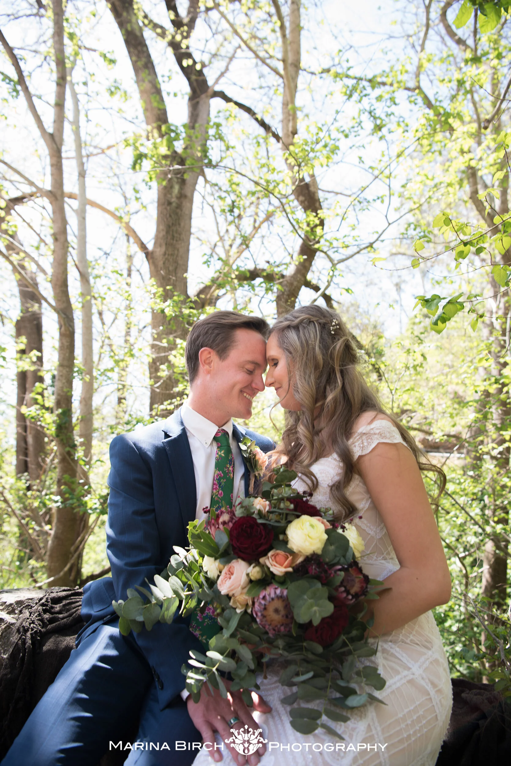 A bride and groom sit close with foreheads touching, smiling in a wooded outdoor setting, holding a large bouquet of flowers.