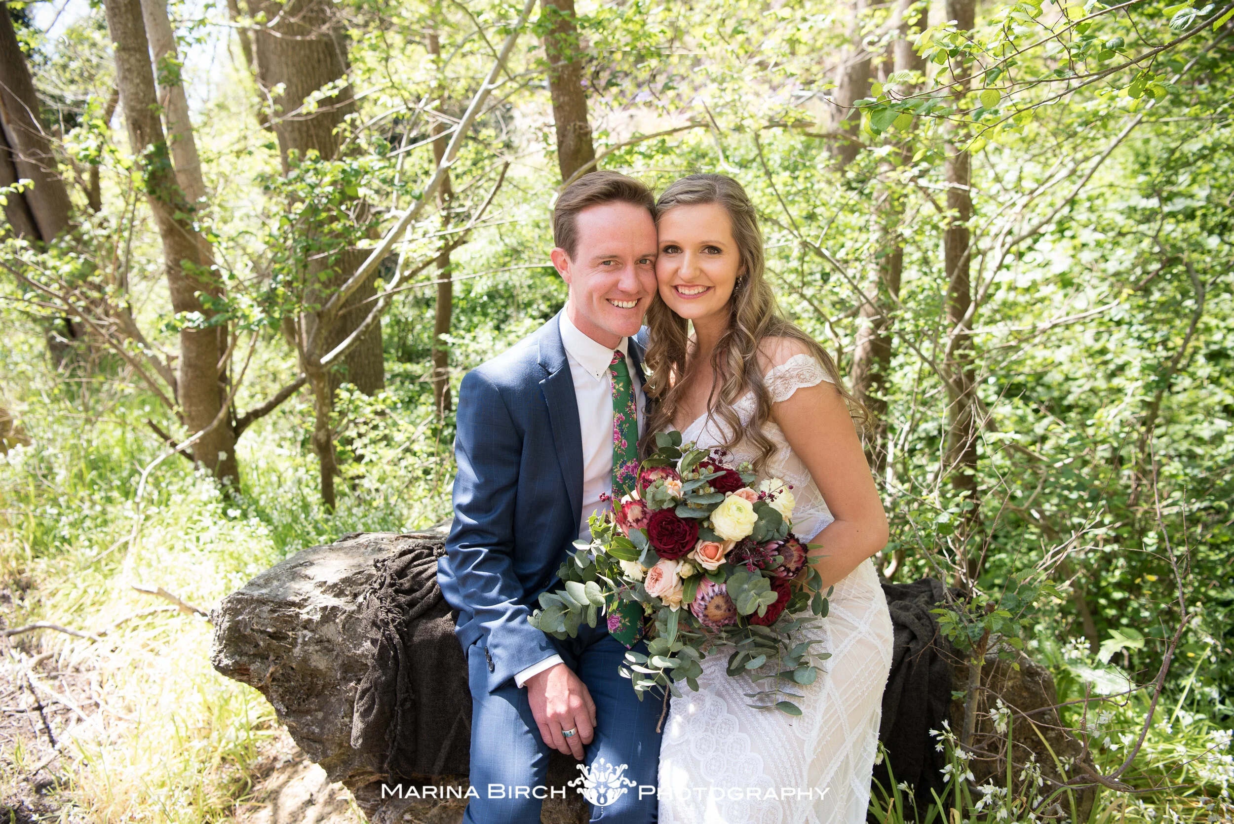 A newly married couple sitting on a large rock in a wooded area, smiling, with the bride holding a bouquet of flowers. The groom is wearing a blue suit, and the bride is in a white lace wedding dress.