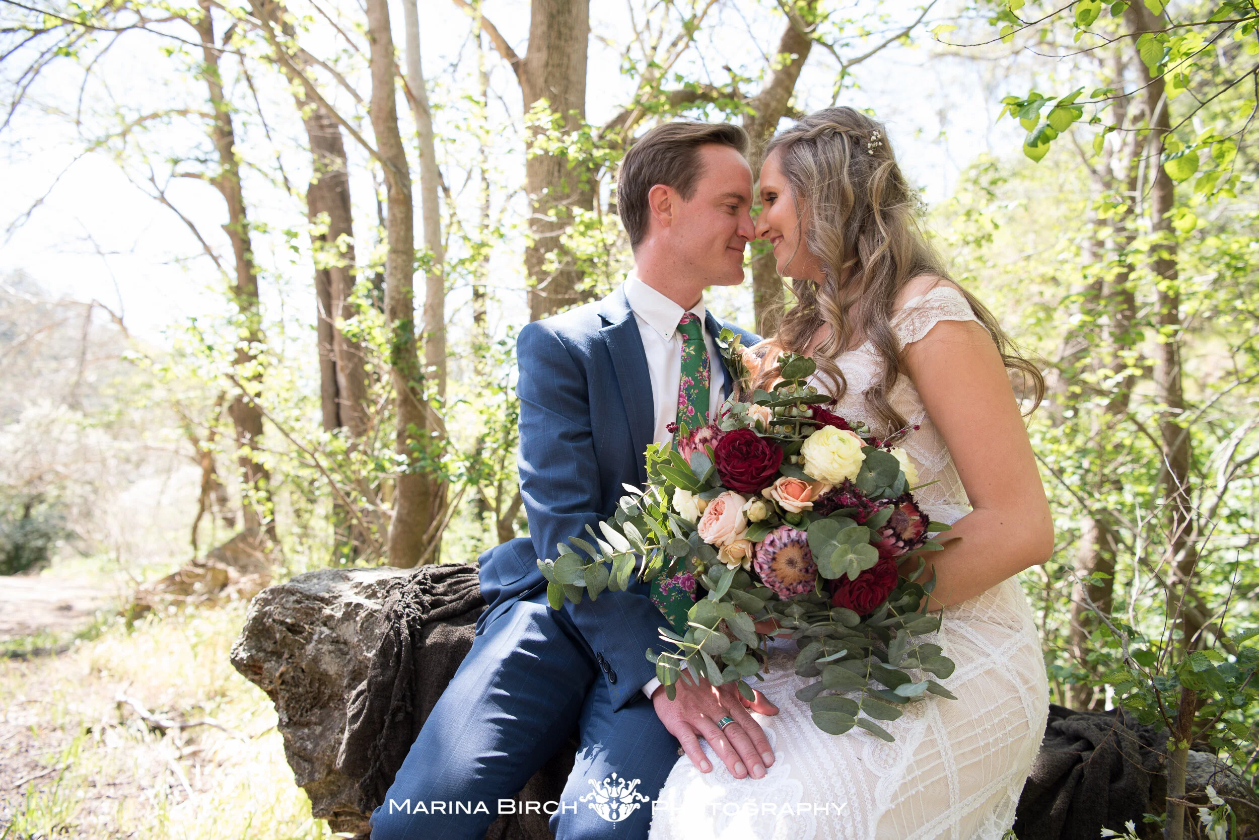 A bride and groom sit closely together outside in a wooded area, touching foreheads and holding a large bouquet of flowers.