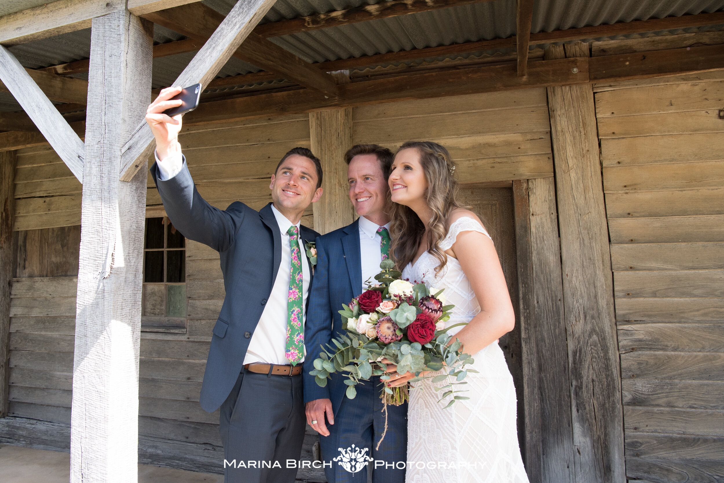 Three people taking a selfie at a wedding, with the bride in a white dress holding a bouquet of flowers. The two men in suits stand beside her, smiling.