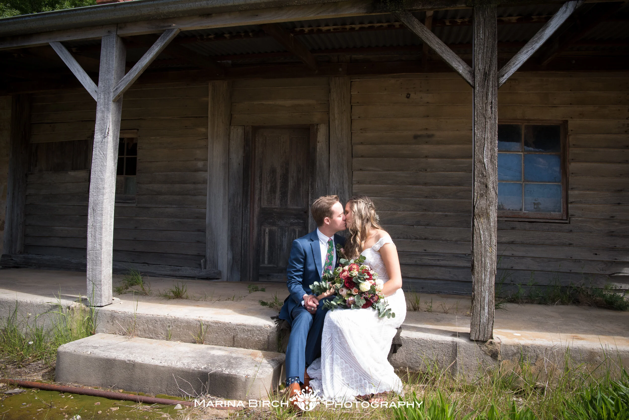 A bride and groom sit on stone steps in front of an old wooden building, sharing a kiss. The bride wears a white lace wedding dress and holds a bouquet of pink, white, and red flowers. The groom is dressed in a blue suit with a floral tie.