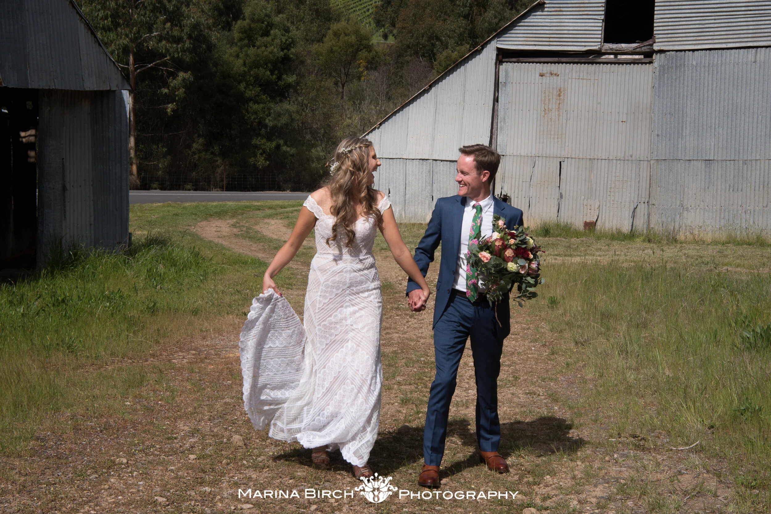 A bride and groom walking hand-in-hand outdoors, smiling at each other, with the bride holding her dress and the groom carrying a bouquet of flowers, in front of a rustic barn and green trees.