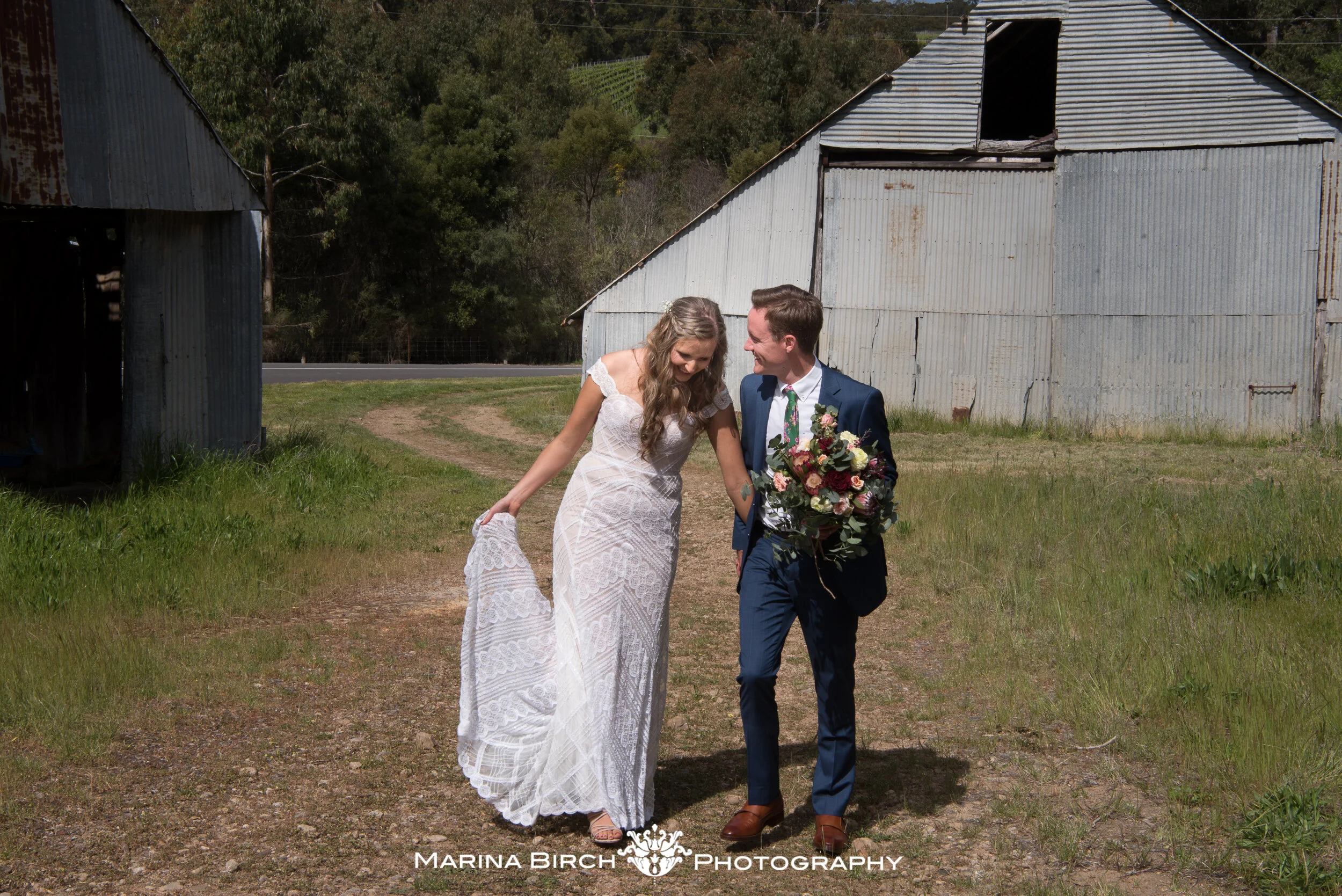 A bride and groom walking on a dirt path beside rustic, weathered barns. The bride is wearing a white lace wedding dress, and the groom is dressed in a dark blue suit, holding a large bouquet of flowers. They are smiling and looking at each other.
