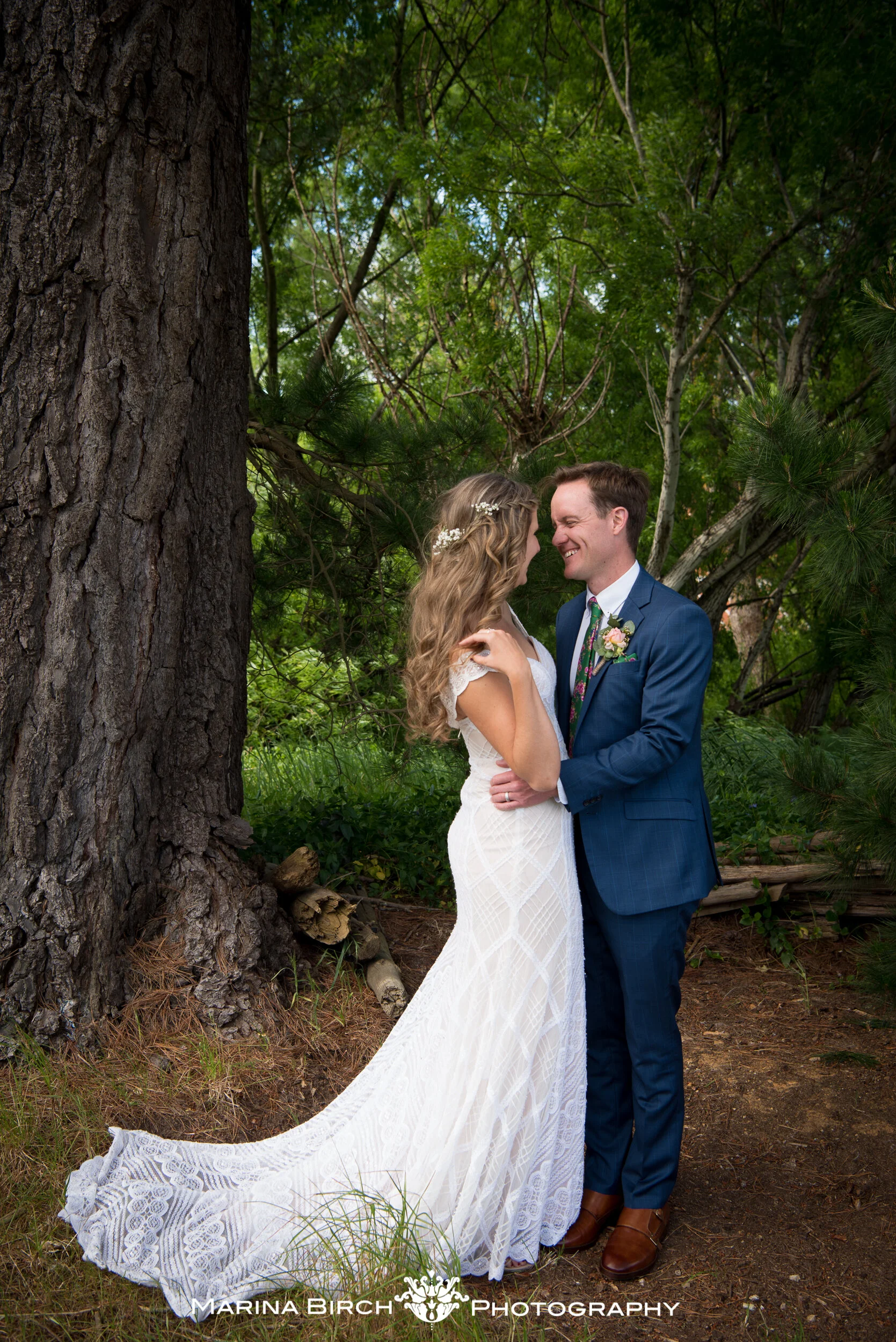 A bride and groom smiling and embracing outdoors on their wedding day, standing near a large tree in a forested area.