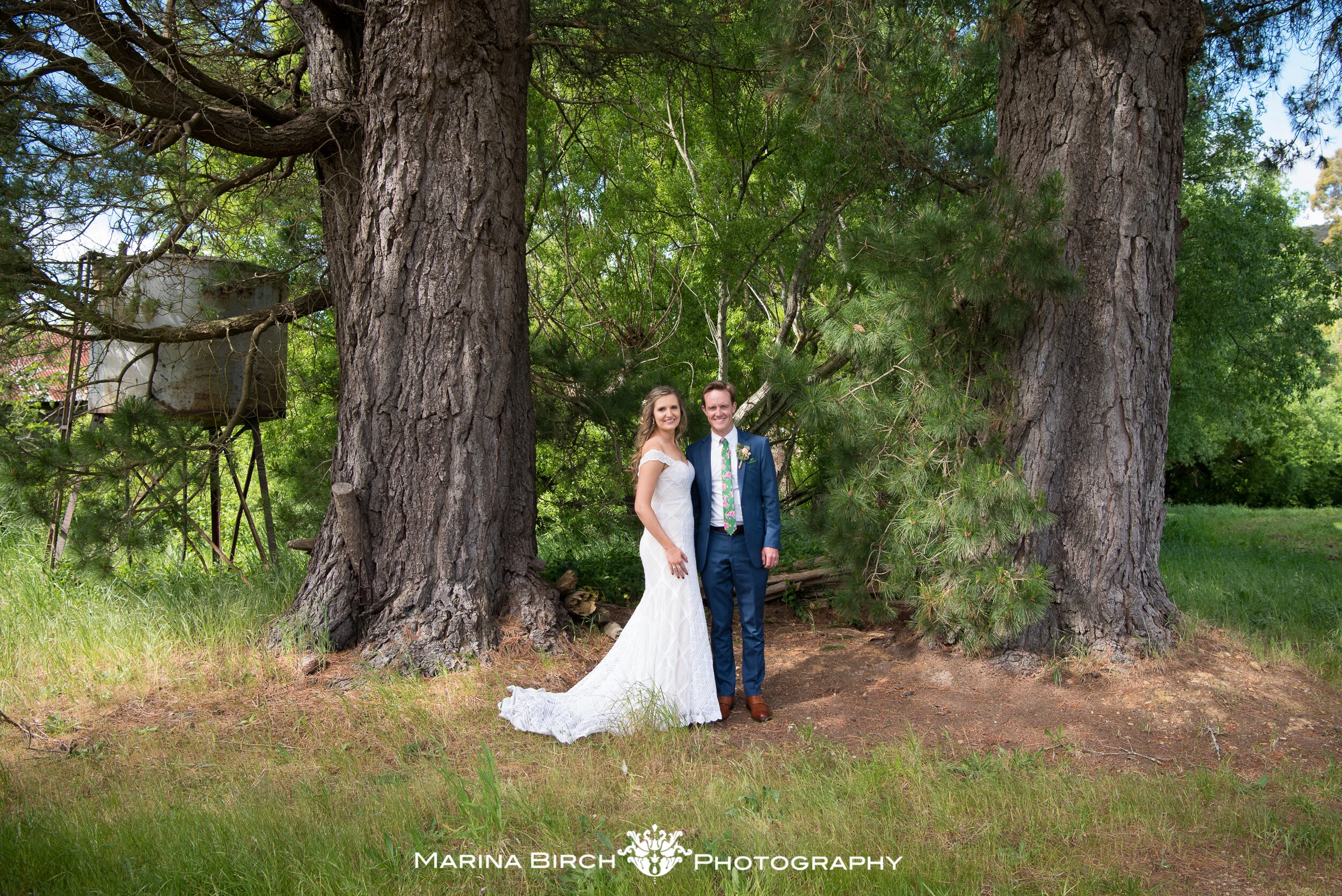 A bride and groom standing together outdoors near two large trees, smiling, during their wedding photos.