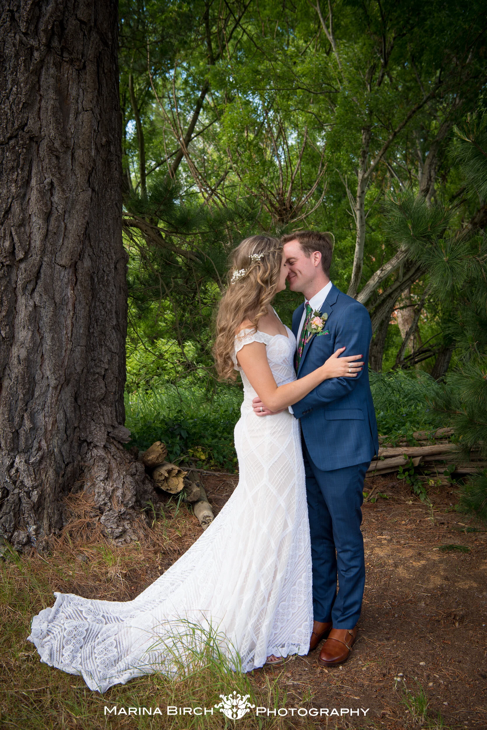 A bride and groom embracing and smiling with foreheads touching outdoors in a lush green forest. The bride is wearing a white lace gown with a train, and the groom is in a blue suit with a floral boutonniere.