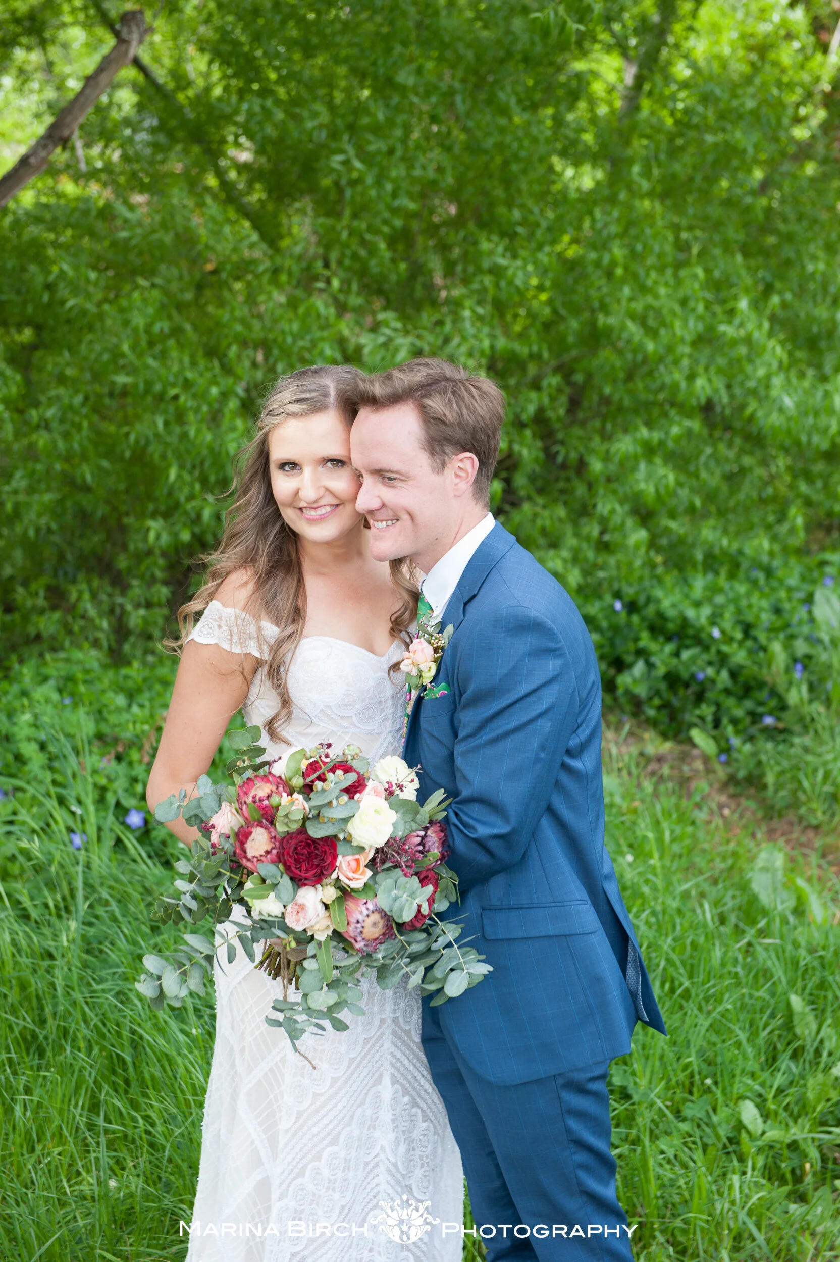 A bride and groom standing close outdoors, smiling, with the bride holding a large bouquet of flowers, surrounded by green trees and grass.