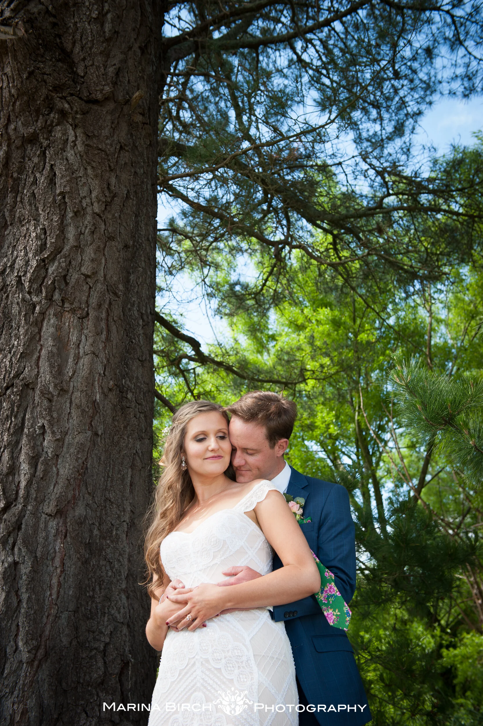 A bride and groom embracing outdoors near a large tree, with greenery and blue sky in the background, during a wedding photoshoot.