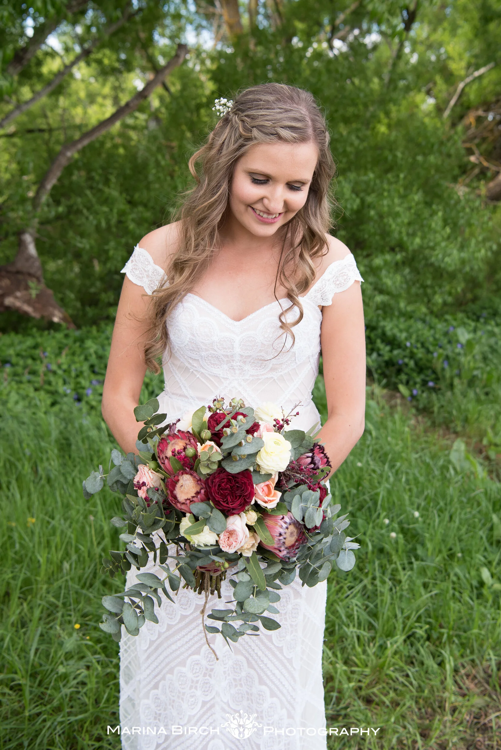 A smiling woman in a white lace dress holding a large bouquet of red, white, and pink flowers, standing outdoors in a green grassy area with trees in the background.