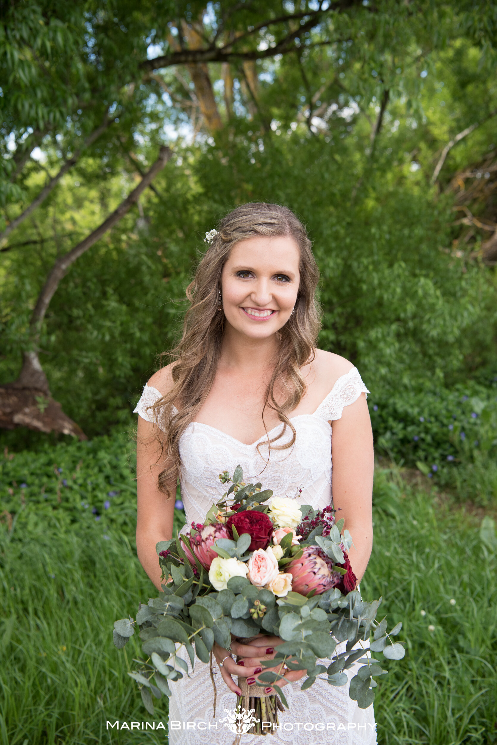 A smiling bride in a white lace dress holding a bouquet of flowers, standing outdoors in a green garden with trees.