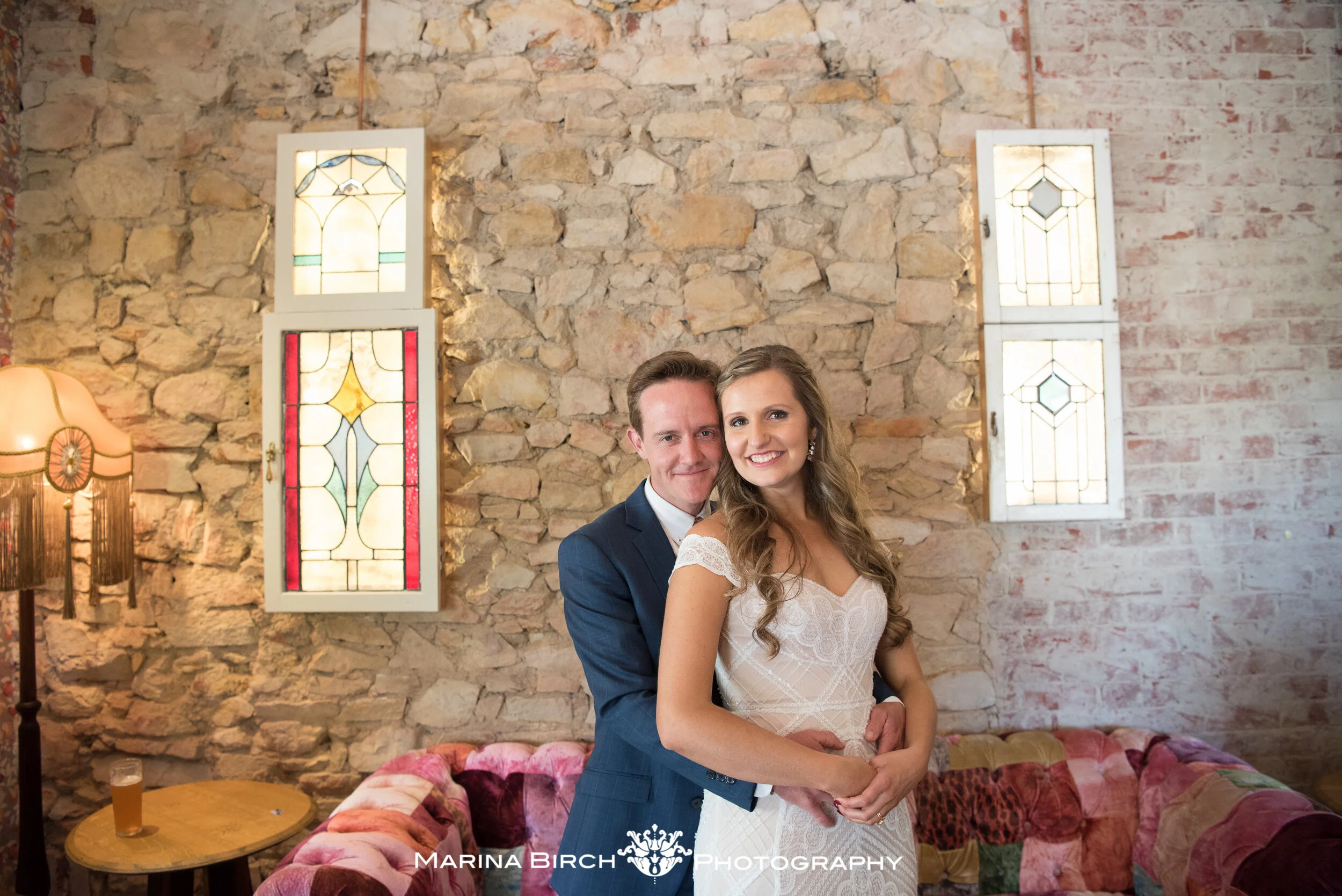 A bride and groom posing together, standing in front of a rustic brick and stone wall with stained glass windows, in a cozy indoor setting with a velvet patchwork sofa and a side table holding a drink.