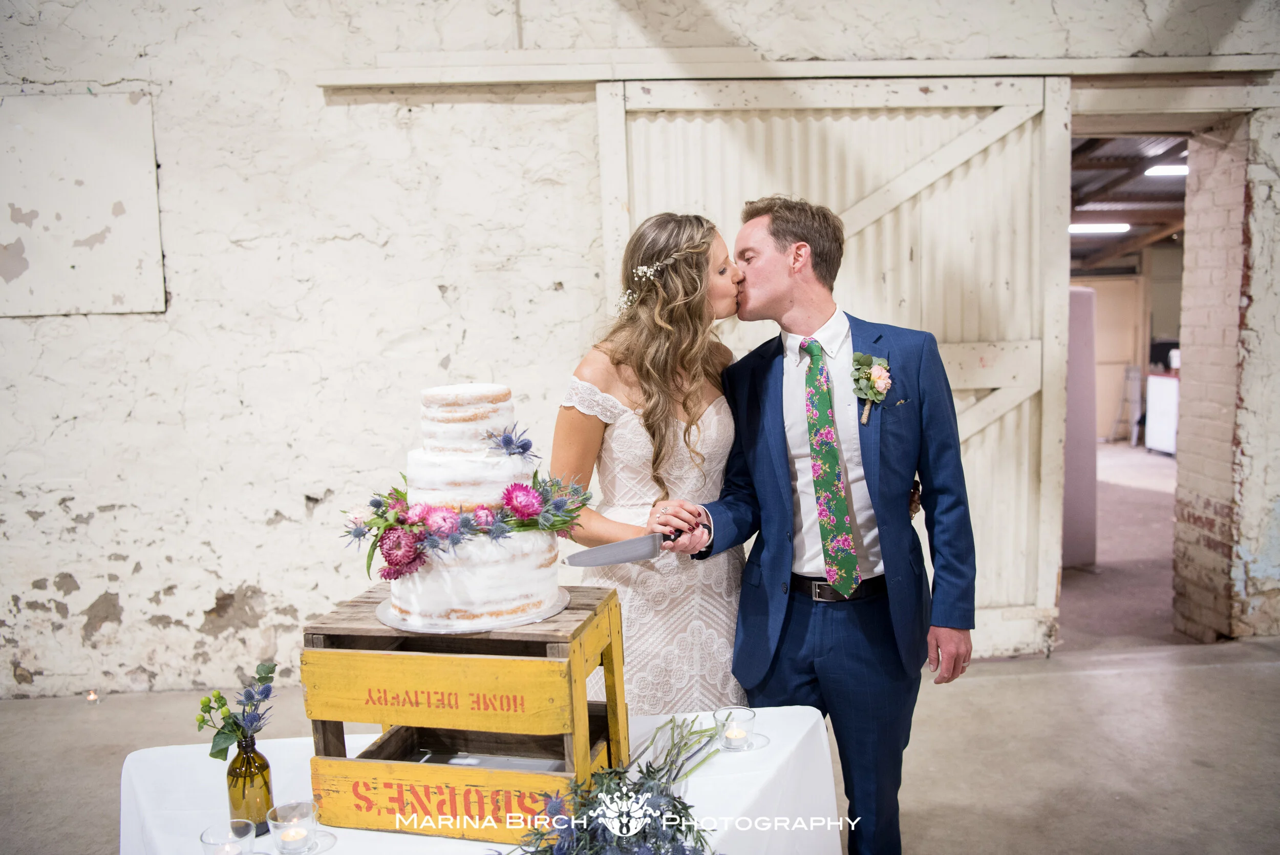 Bride and groom sharing a kiss at their wedding reception, near a decorated wedding cake, in a rustic indoor setting.